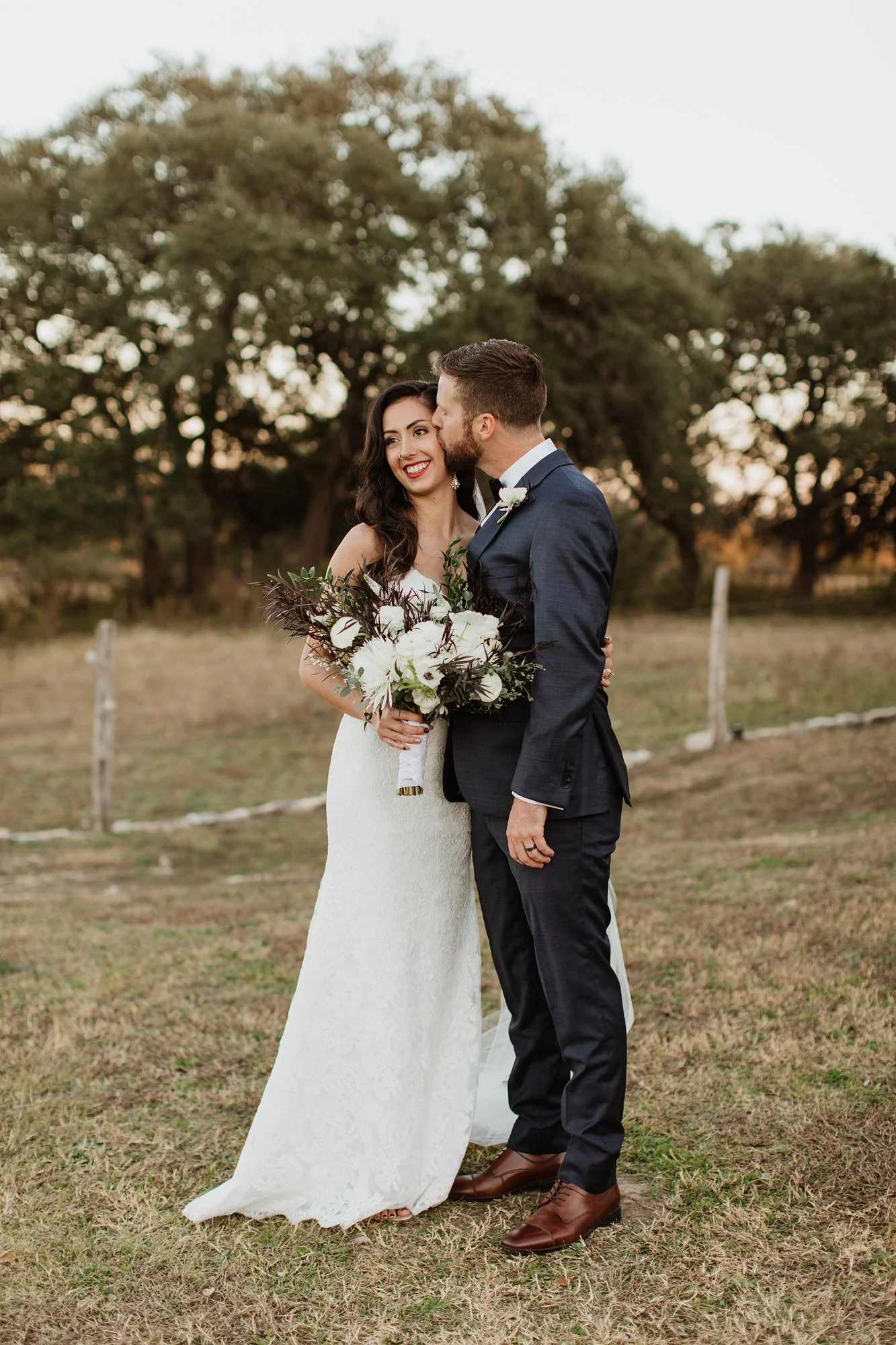 A bride and groom sharing a moment outdoors during sunset; the bride in a white lace gown holding a bouquet, smiling; the groom in a dark suit with a boutonniere, kissing her on the cheek, with trees and open field in the background.