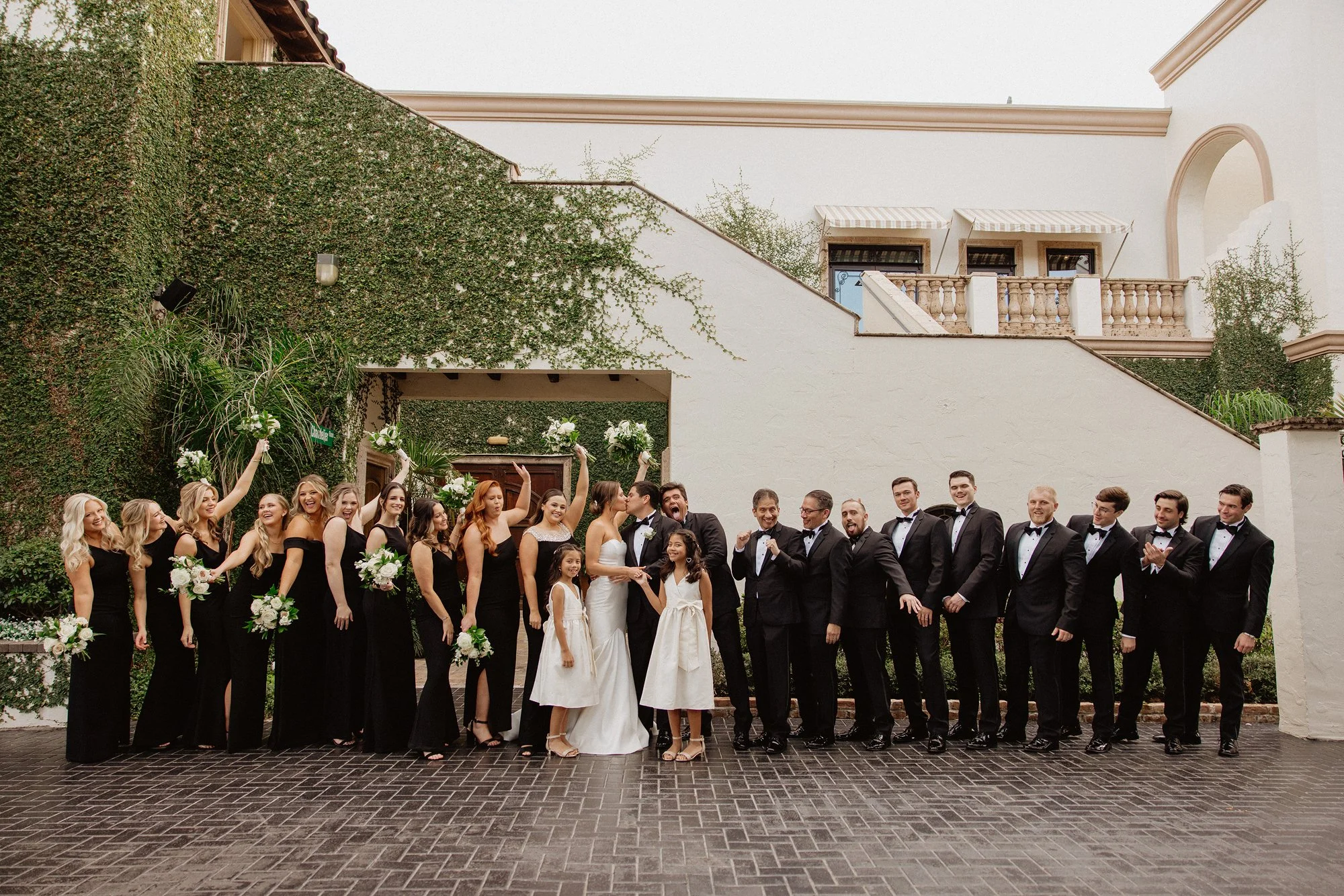 A group of wedding celebration attendees posing outdoors. Bridesmaids and groomsmen, dressed in black and tuxedos, stand in front of a white building with greenery on the wall. The bride and groom are in the center, surrounded by smiling guests holdi