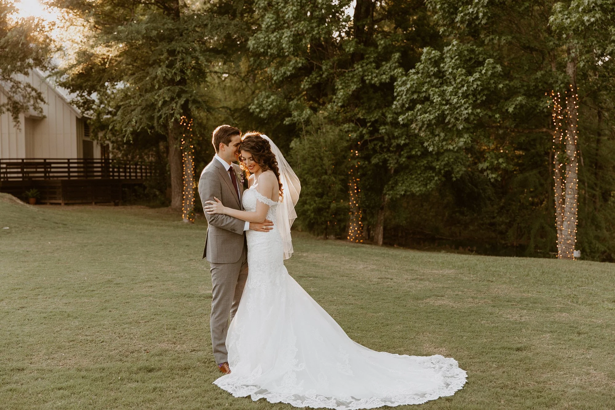 A bride and groom embrace outdoors, with the bride in a white wedding gown with lace details and a veil, and the groom in a gray suit with a maroon tie. They are standing on a grassy area surrounded by large trees decorated with small string lights, 