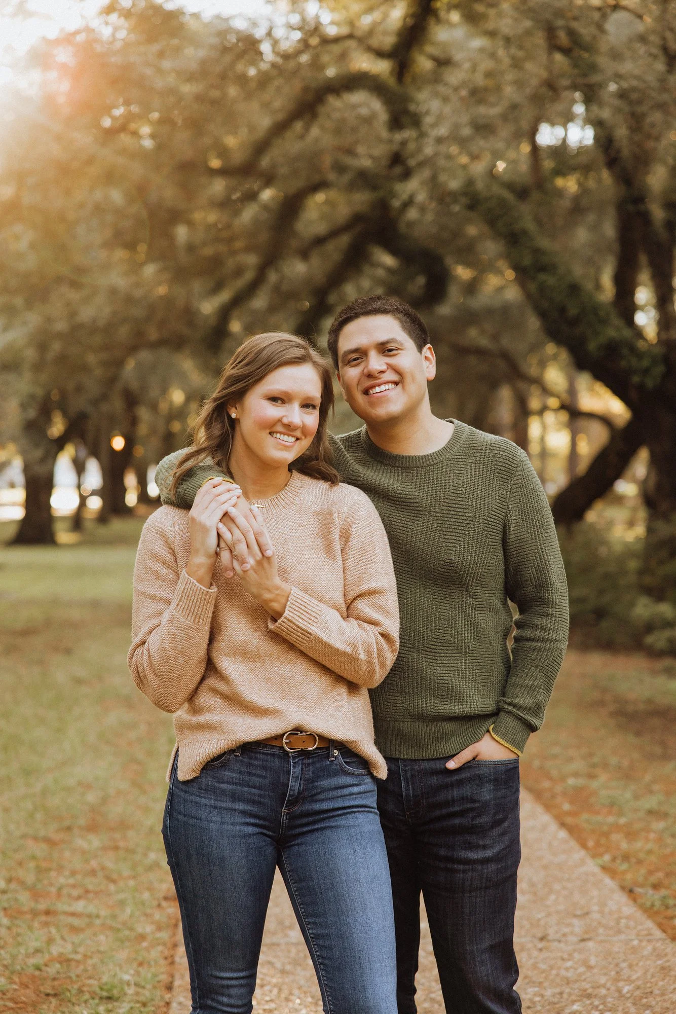 A young couple standing outdoors on a tree-lined path during fall, smiling and embracing each other.