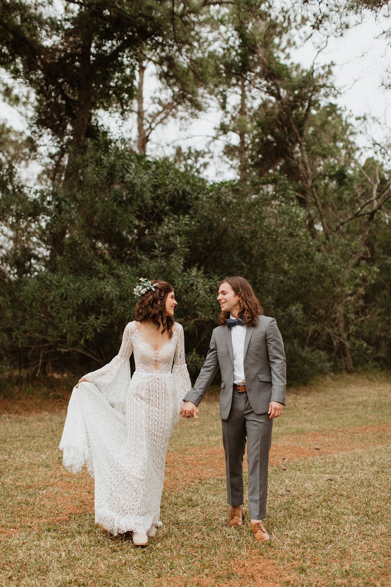 A bride and groom walking hand in hand outdoors, surrounded by trees and grass, smiling at each other.