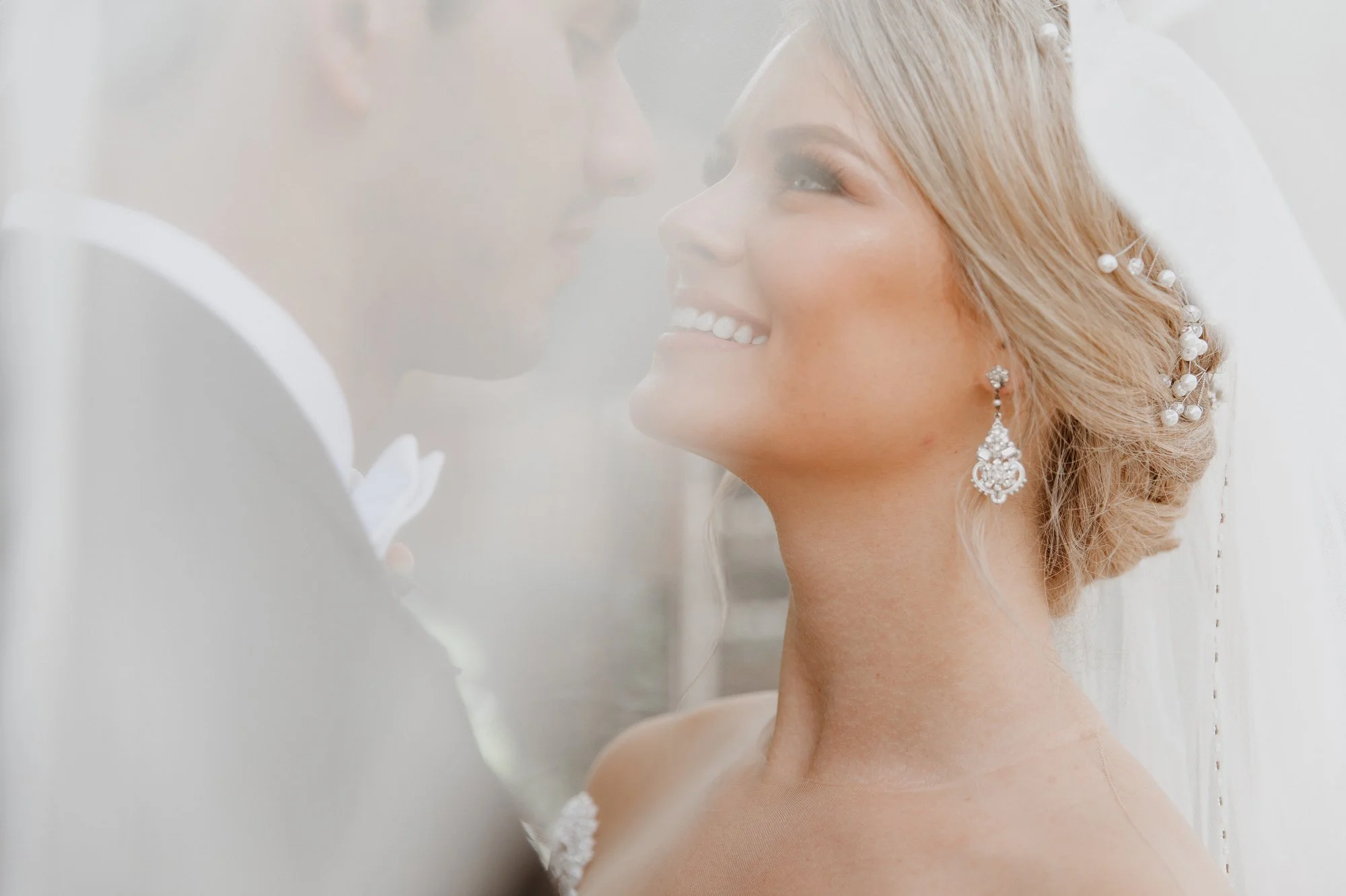 A bride and groom close together, smiling, with the bride wearing pearl earrings and hair accessories, and the groom in a tuxedo.