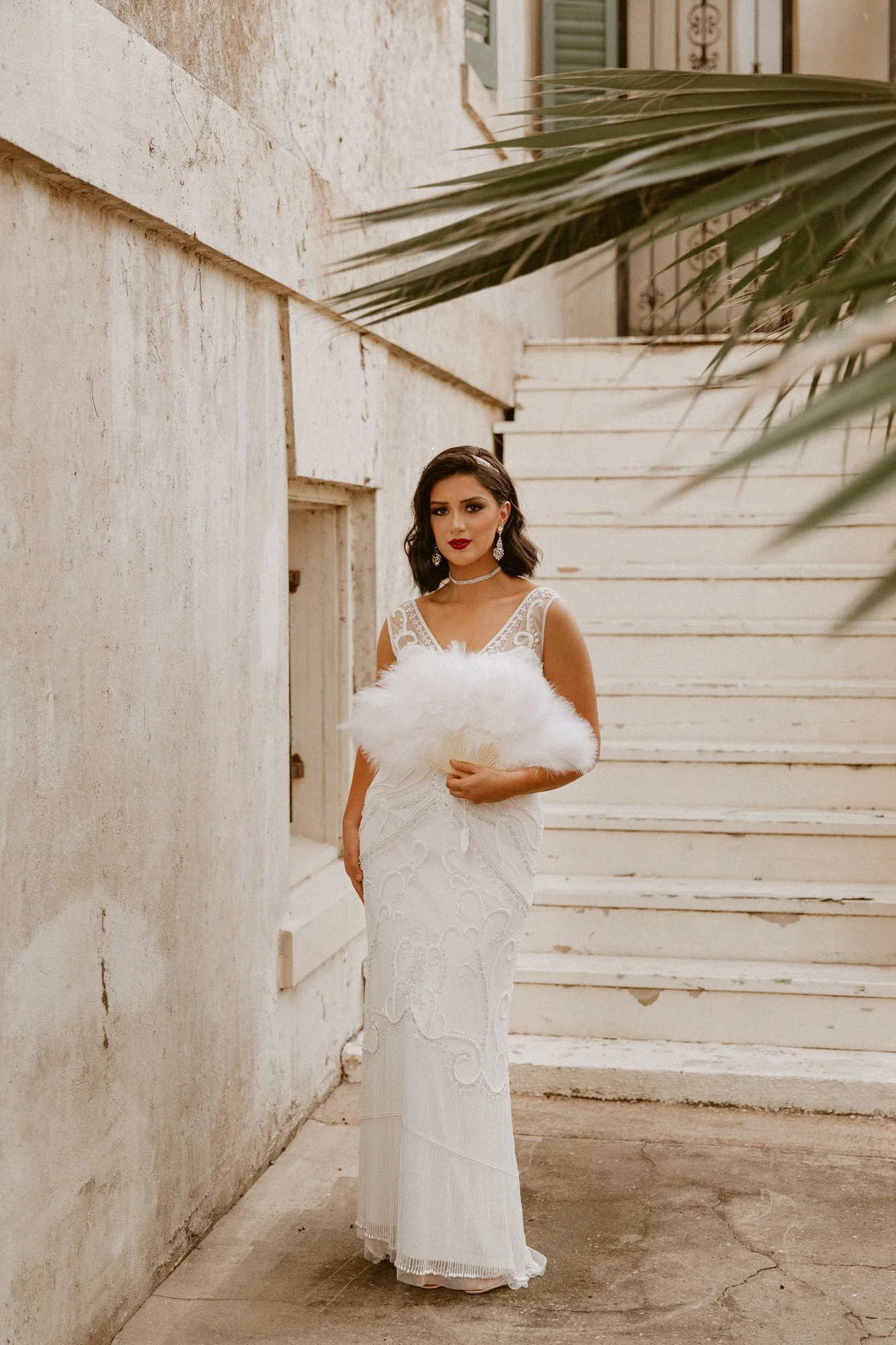 A woman in a white wedding dress holding a feathered fan, standing outdoors by a textured beige wall and white steps, with palm leaves partially visible in the upper right corner.