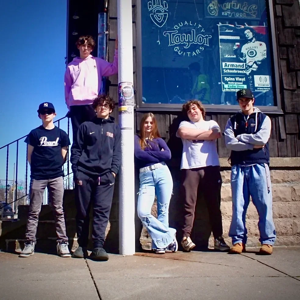 Group of six young people standing outdoors in front of a building with a blue window, with some crossing arms and looking at the camera, under a clear blue sky.