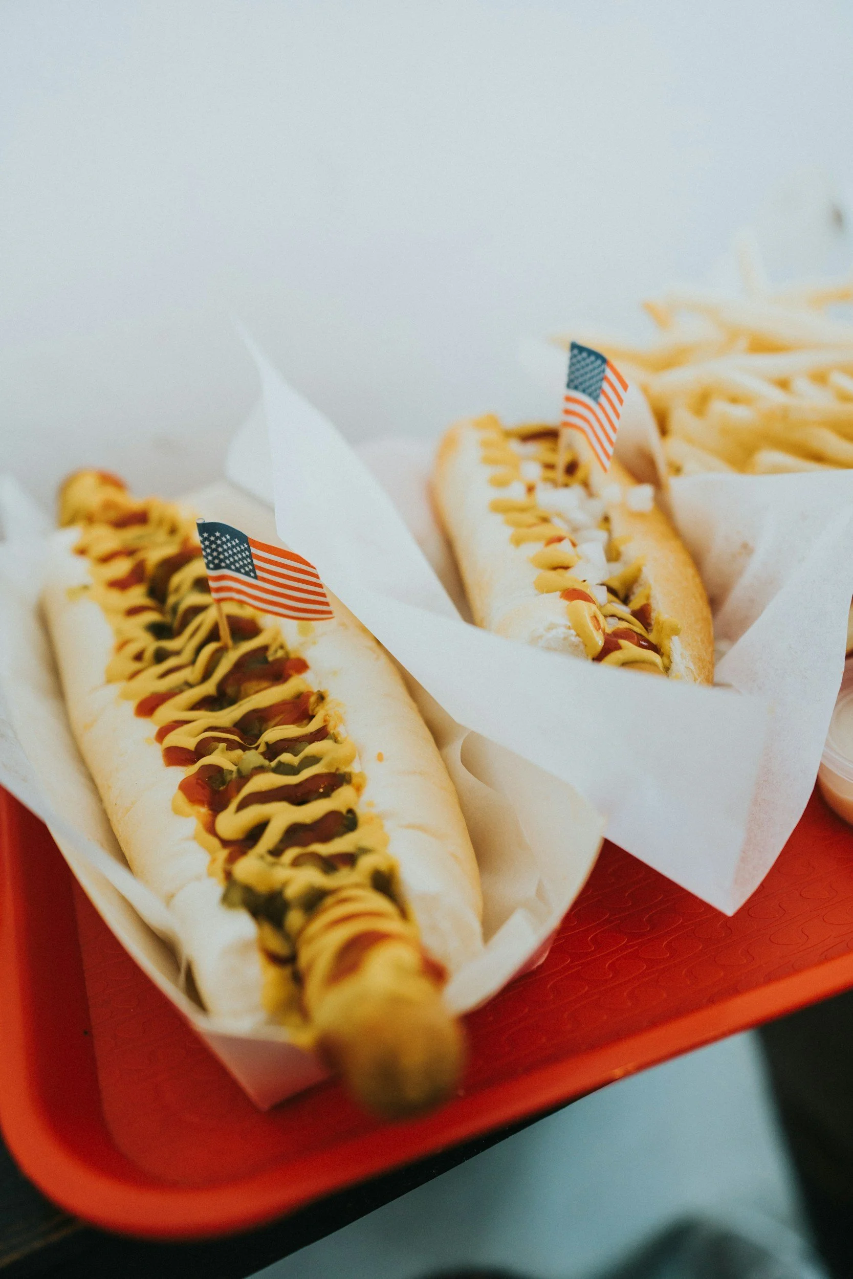 Two hot dogs in buns topped with mustard, ketchup, relish, and mayonnaise, each with small American flags on toothpicks placed in them, served on a white tray with a red border.