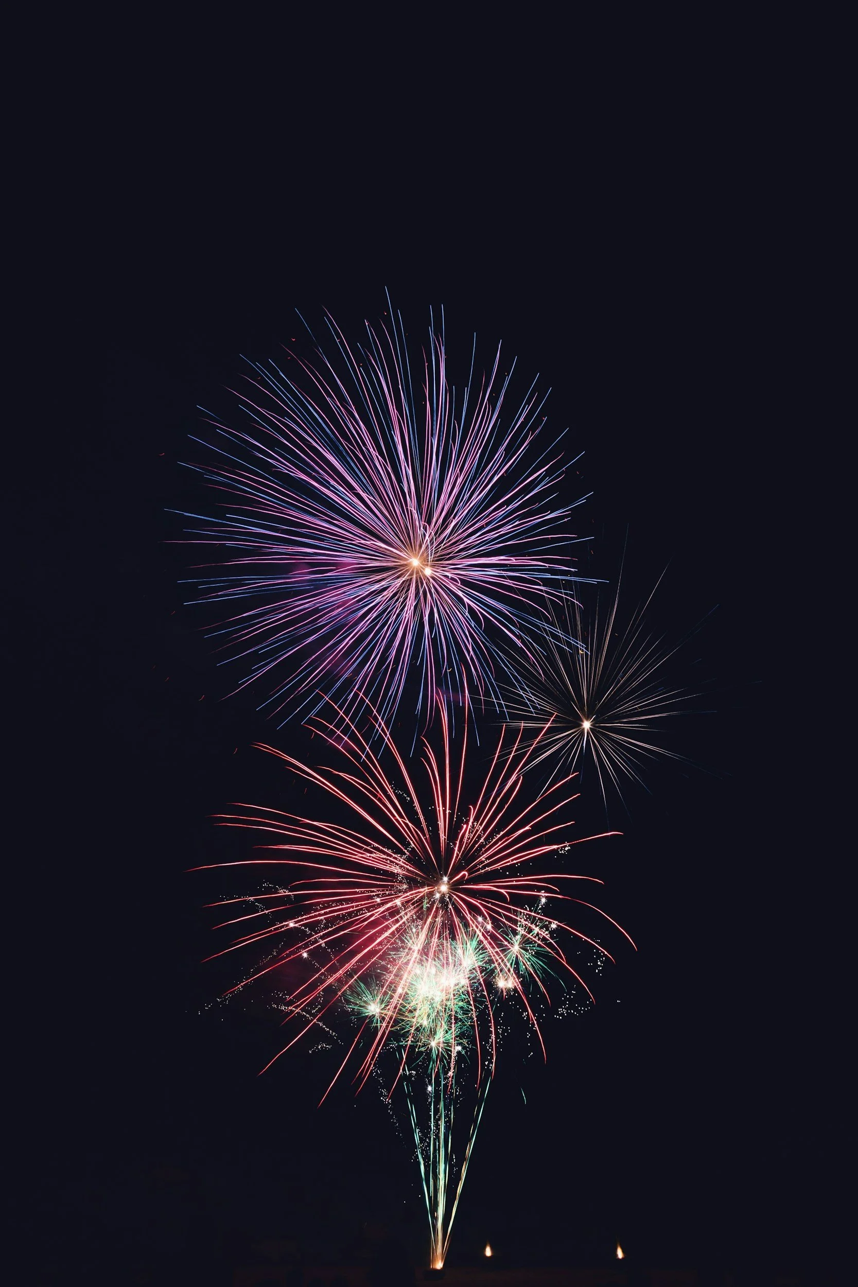 A series of colorful fireworks exploding in the night sky, creating bright streaks of pink, purple, red, green, and blue against the dark background.
