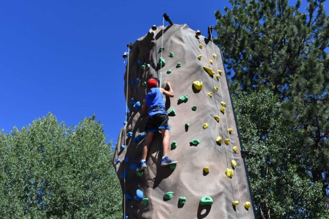 Child wearing a red hat climbing an outdoor rock wall with colorful holds under a clear blue sky