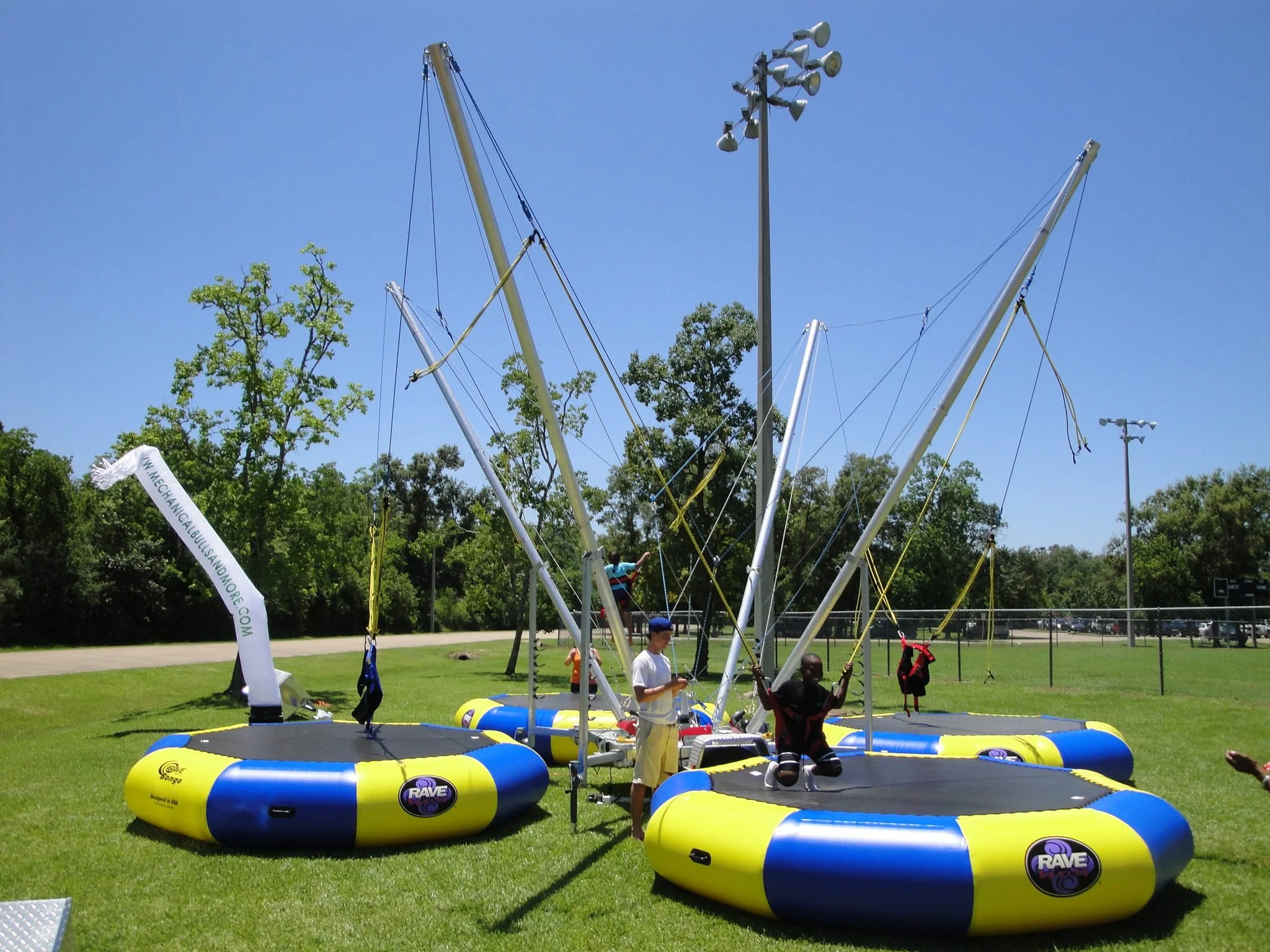 People prepare for a trampoline park on a field with inflatable barriers, trees, and a clear blue sky.