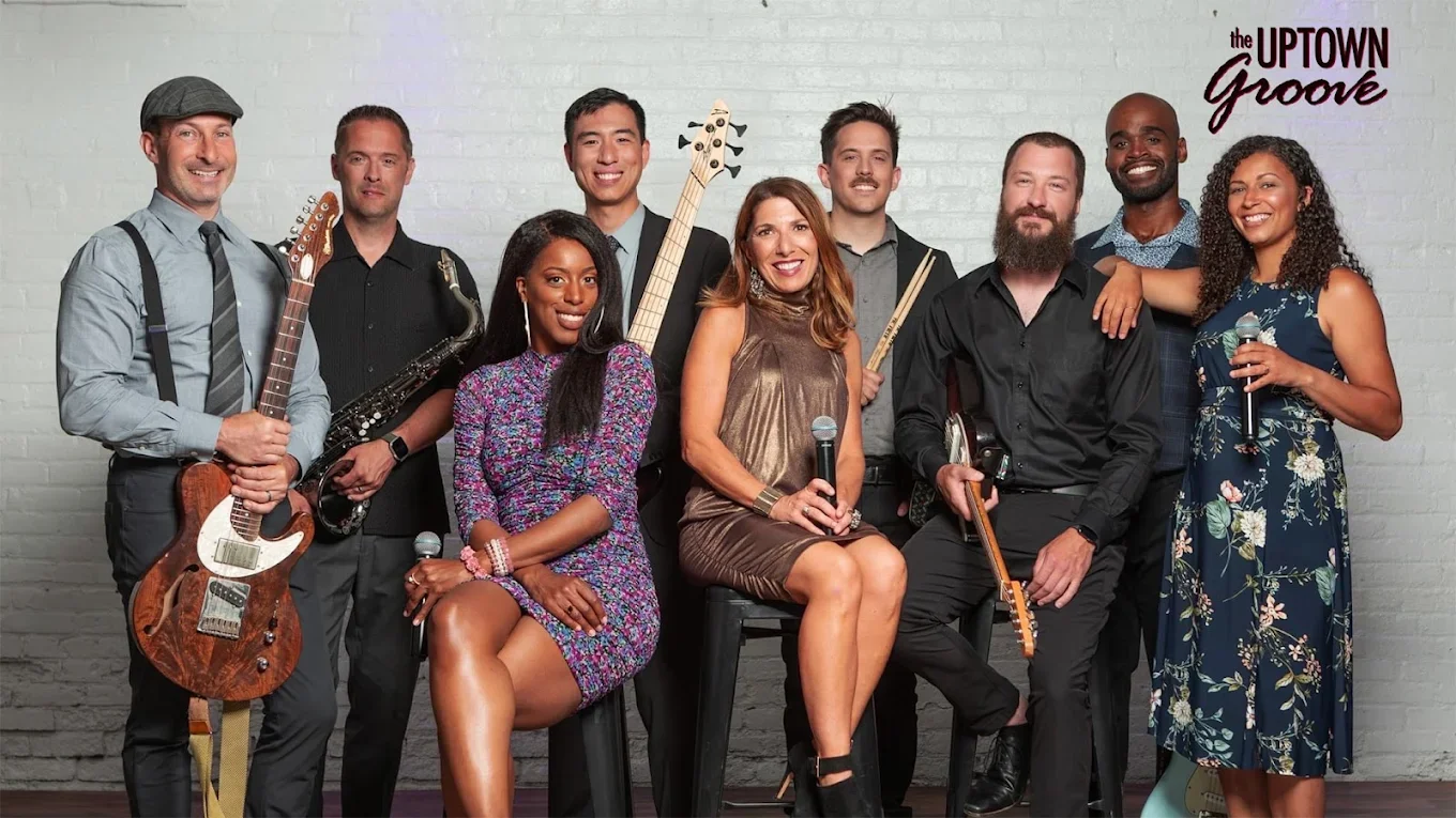A diverse group of nine musicians posing together against a white brick wall, some holding musical instruments and microphones. The words 'the UPTOWN Groove' appear in the upper right corner.