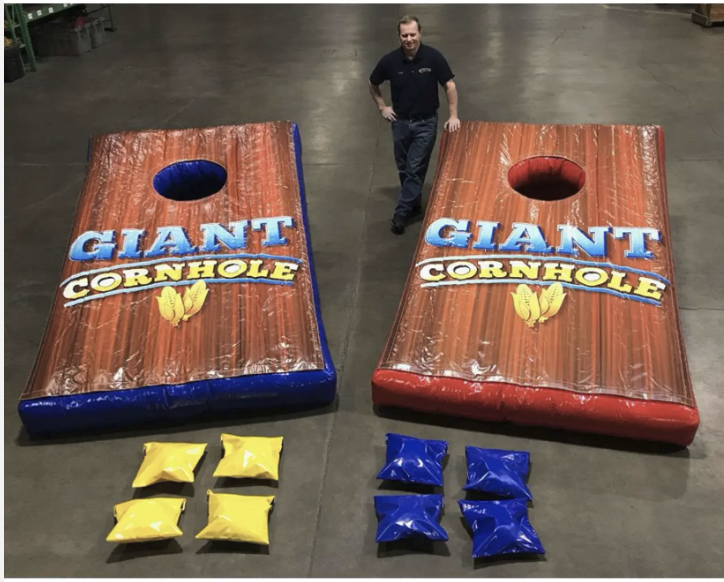 Two giant cornhole game boards with the logo "Giant Cornhole" on them, one with yellow bean bags and one with blue bean bags, inside a warehouse or indoor space with a person standing between them.