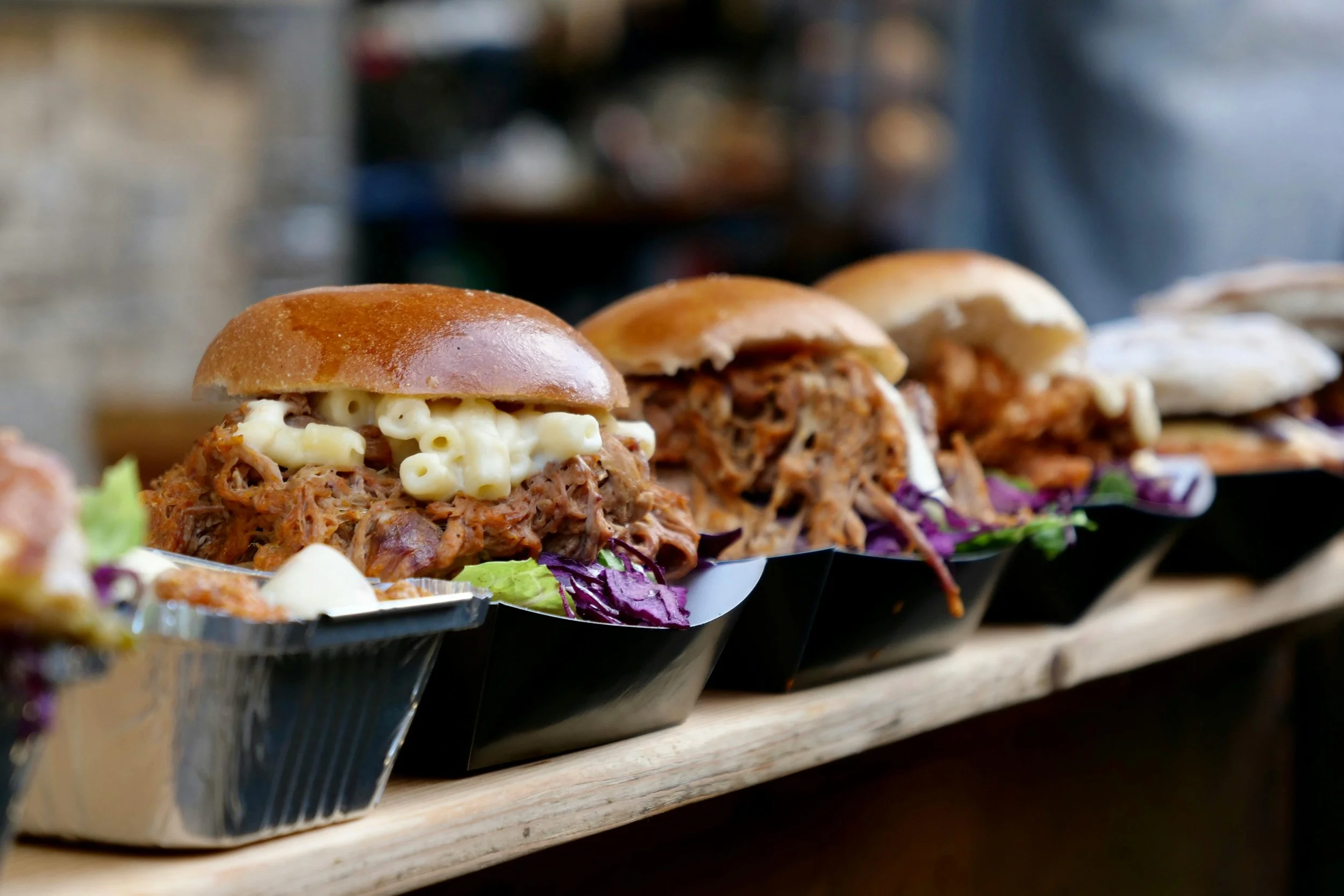 Several pulled pork sandwiches with macaroni and cheese on top, served in black containers on a wooden surface.