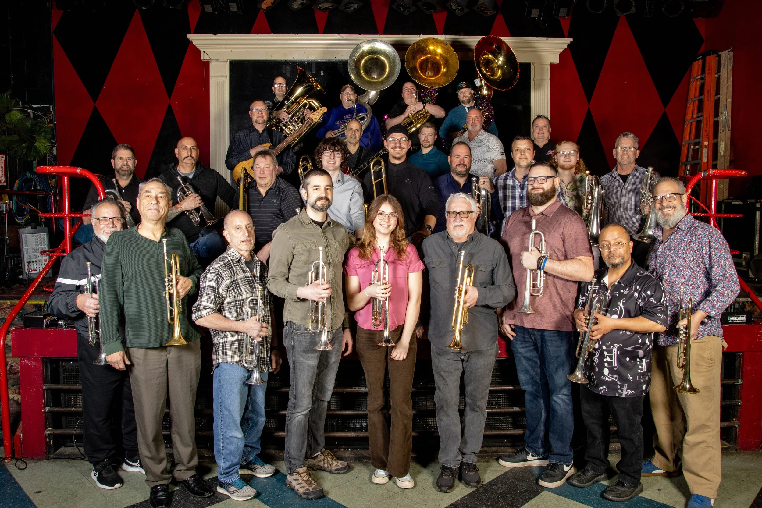A large group of musicians, including trumpet and trombone players, standing in front of a stage with a red and black diamond-patterned wall, some holding their instruments and smiling.