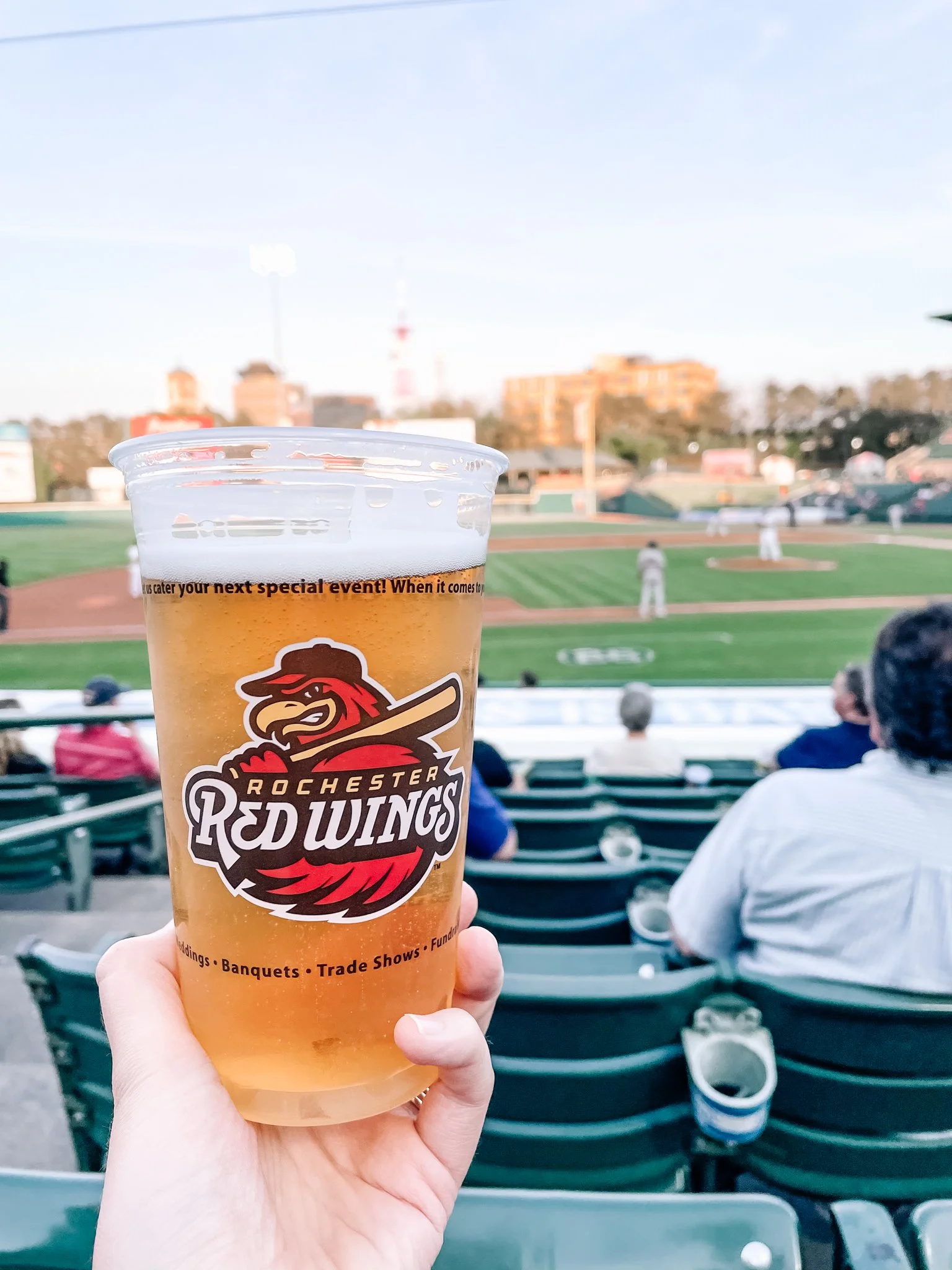 A plastic cup of Rochester Red Wings baseball game beer held up in front of a baseball stadium with players on the field, spectators, and people sitting in the stands visible in the background.