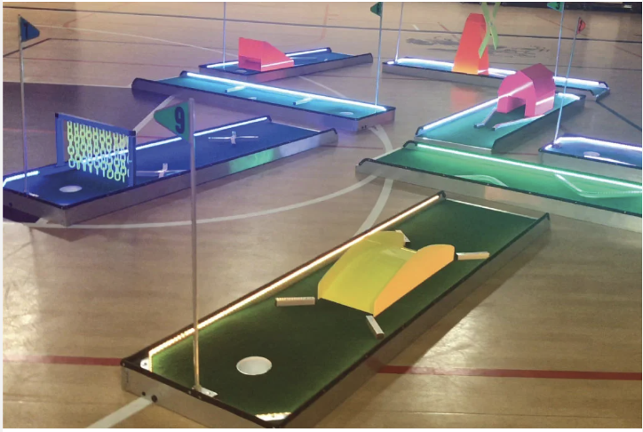 Miniature indoor golf course with multiple colorful holes and flags displaying numbers, illuminated pathways, on a gymnasium floor.