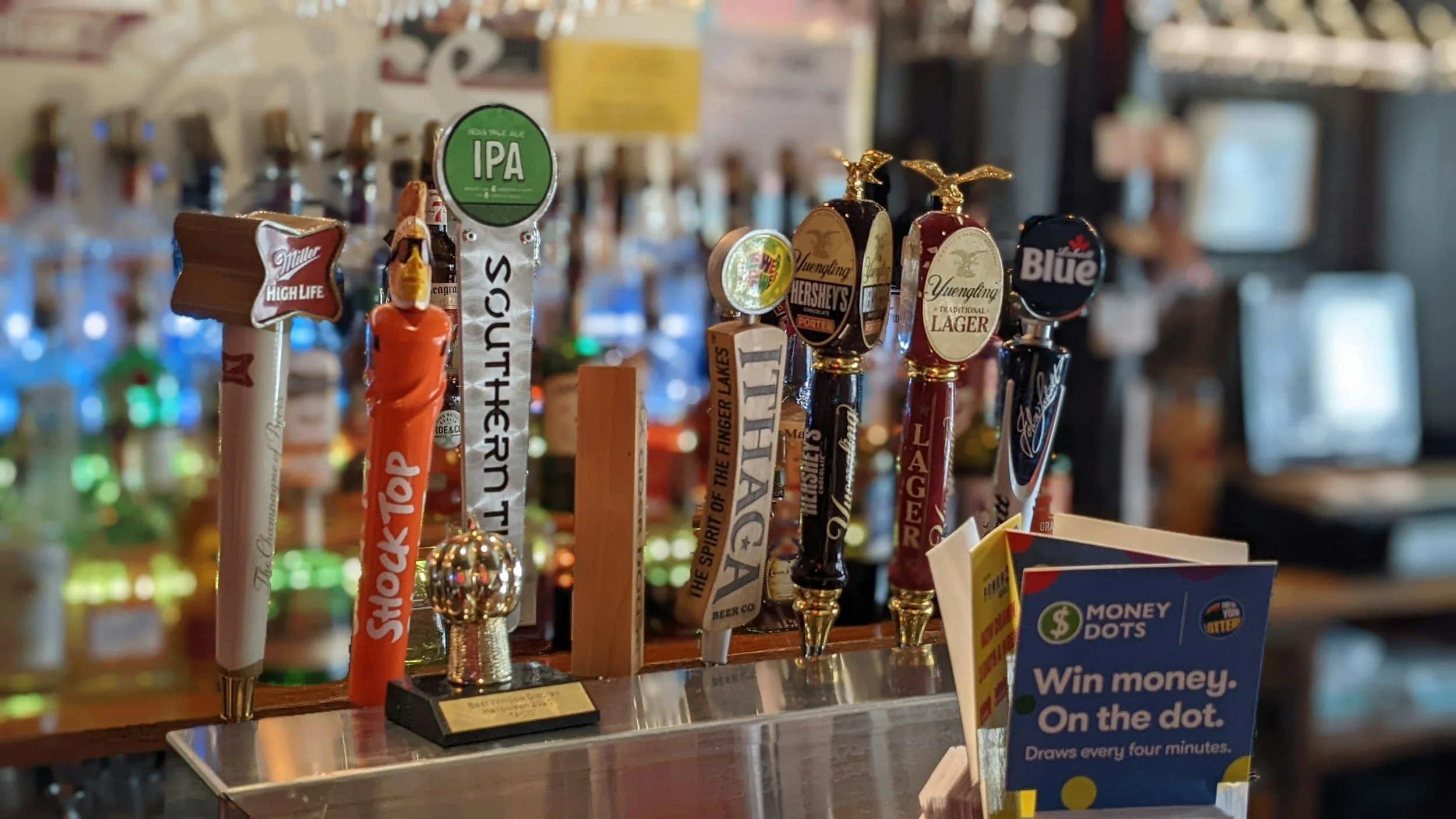 A row of beer tap handles at a bar, featuring various brands and styles, with promotional flyers and a blurry background of liquor bottles.
