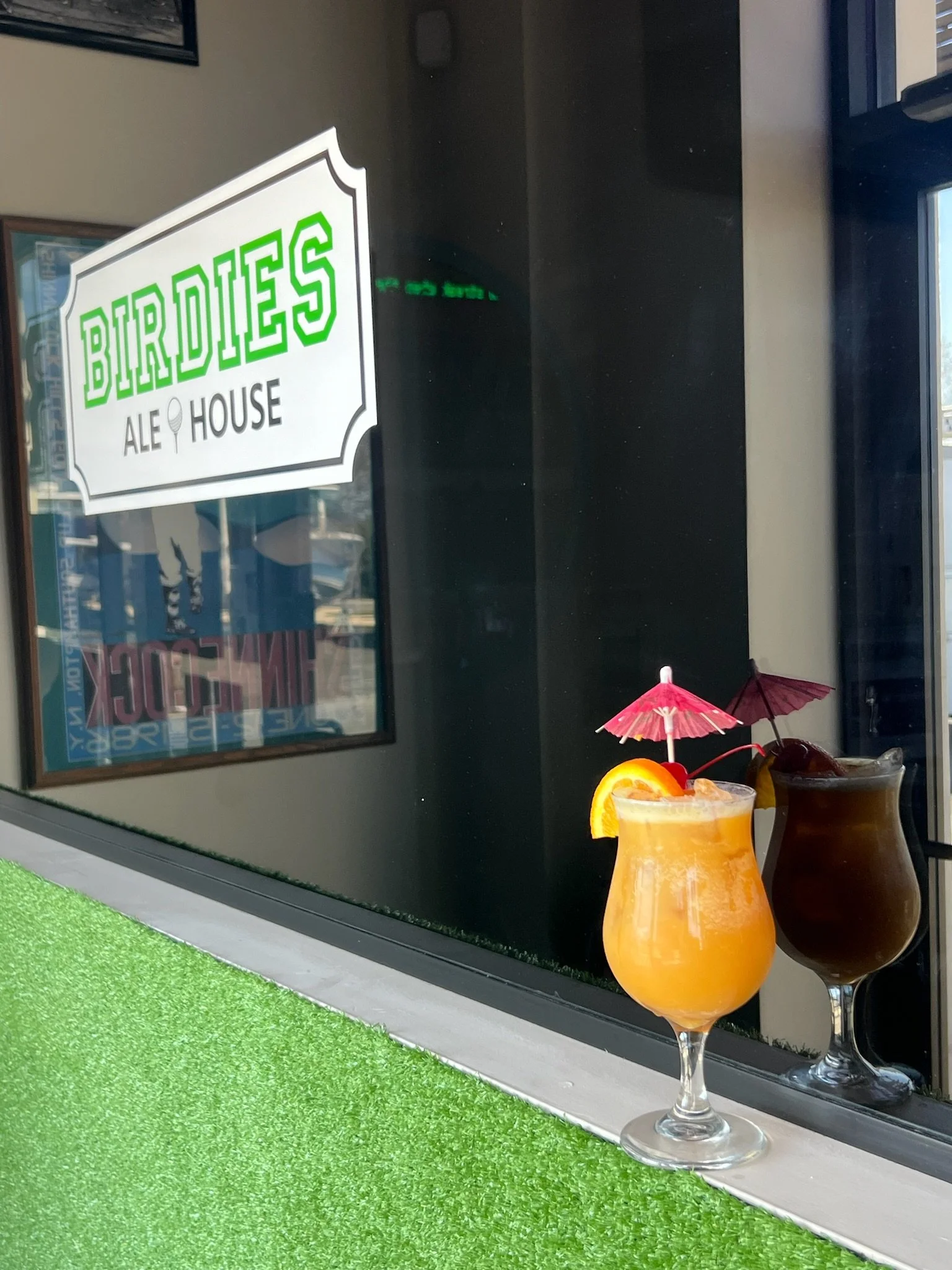 Two tropical drinks with small umbrellas on a green surface outside a bar called 'Birdies Ale House'