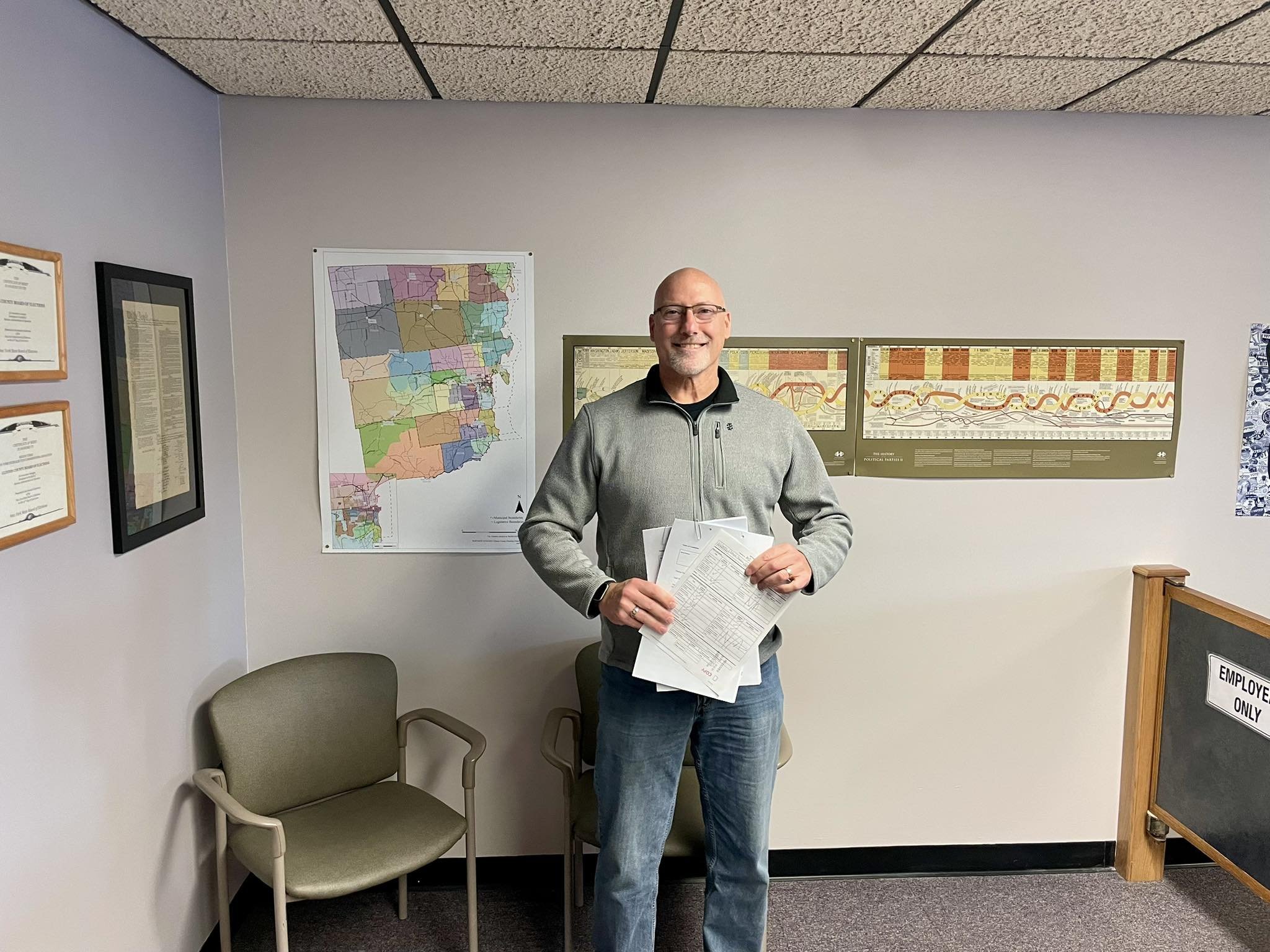 Brent wearing glasses and a gray zip-up jacket standing in an office waiting area with chairs and framed posters on the wall. He is holding papers in his hands.