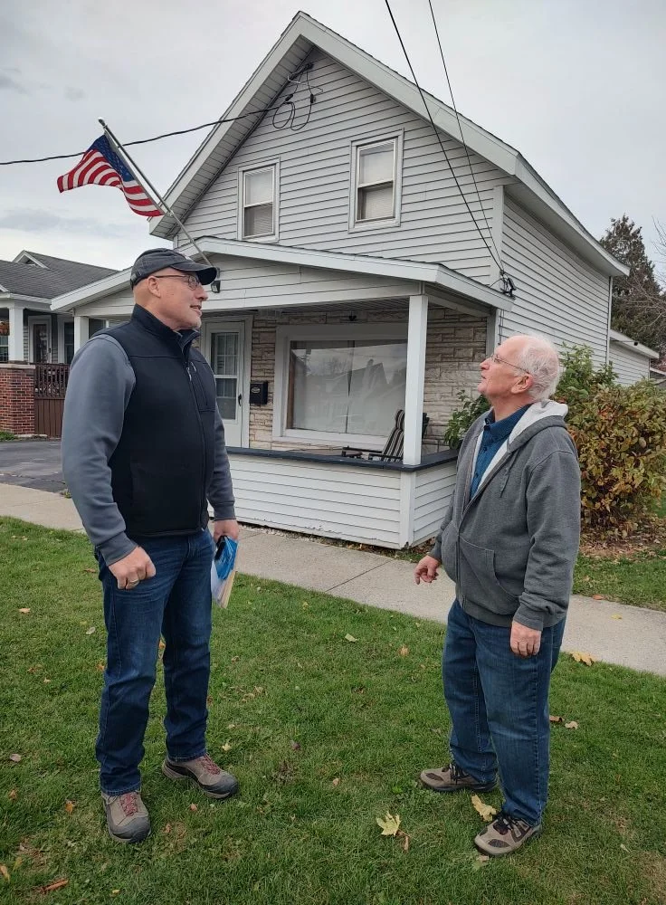 Brent engaged in conversation with Clinton County resident in front of his home. The house is a two-story with white siding, an American flag on a flagpole, and a porch with a large front window.