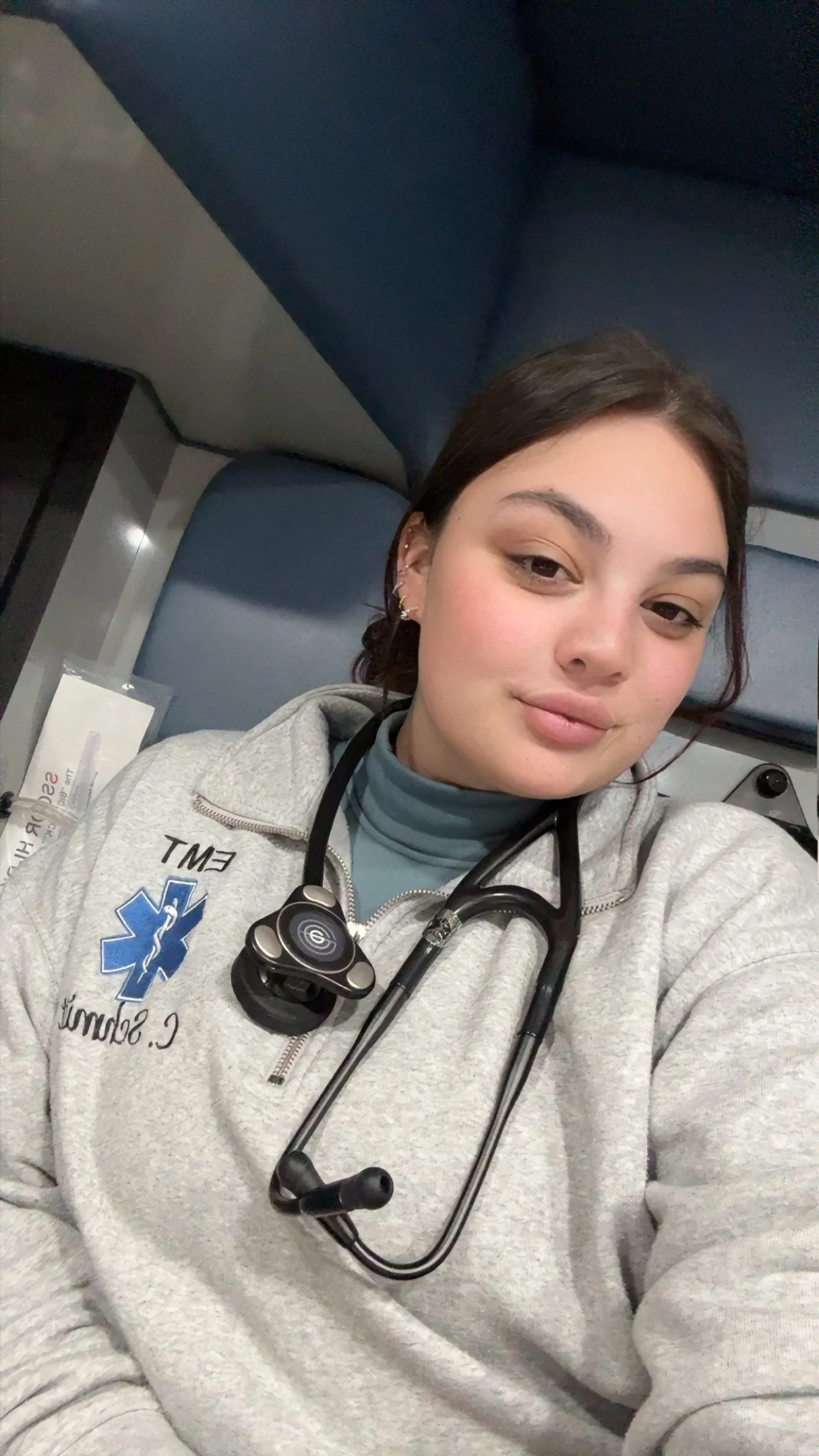 A young woman with dark hair, wearing medical scrubs and a gray zip-up jacket with an EMT patch, sitting on a blue seat in an emergency vehicle or ambulance, with a stethoscope around her neck and a calm expression.