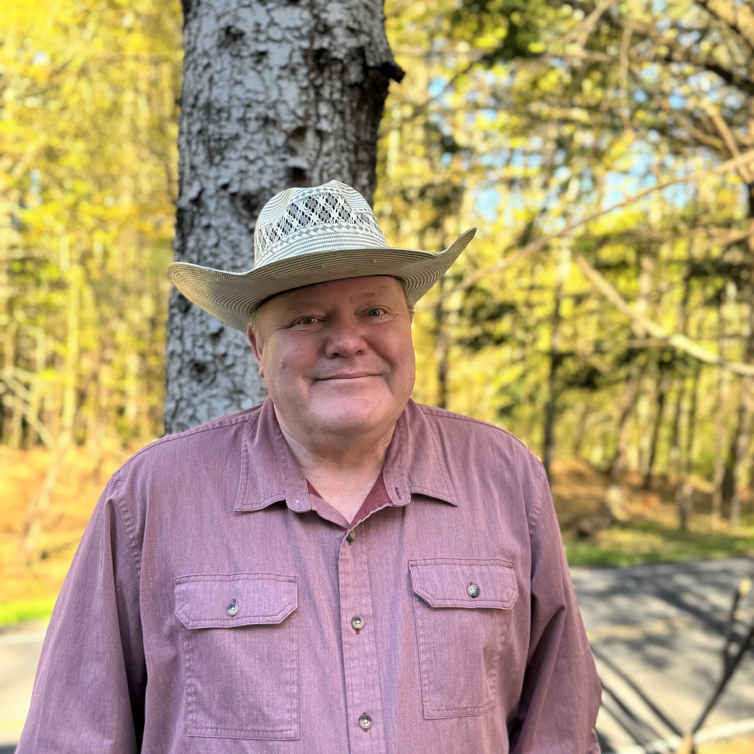Person with fair skin wearing a wide-brimmed patterned hat and a mauve-colored button-up shirt, standing outdoors near a tree with yellow and green foliage in the background.