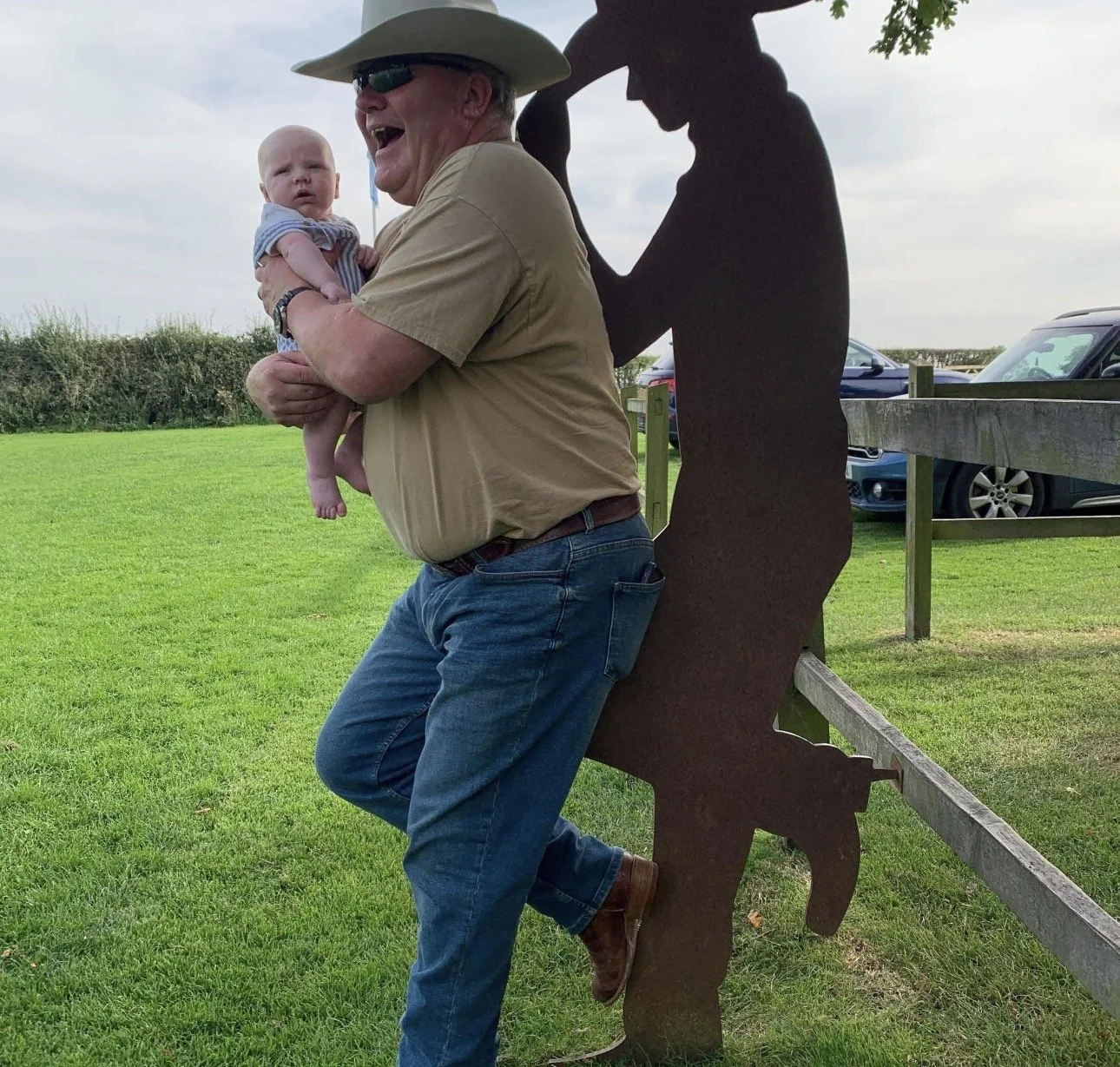 An elderly man with a hat, sunglasses, and a tan shirt is holding a baby with a striped shirt outdoors on a grassy field. They are beside a wooden fence with a silhouette of a person cutout behind them, and some cars are parked in the background.