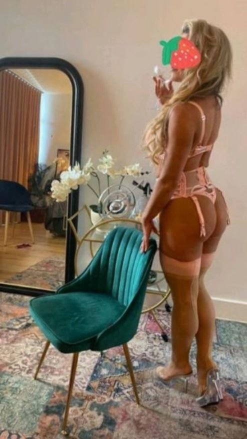A woman with long blonde hair wearing pink lingerie and clear high heels taking a mirror selfie in a room with a green chair, white flowers, and a large mirror reflecting a sitting area.