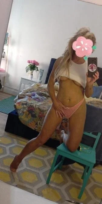 A woman with long blonde hair taking a mirror selfie in a bedroom. She is wearing a white crop top lifted to reveal her midriff and red and white striped underwear.