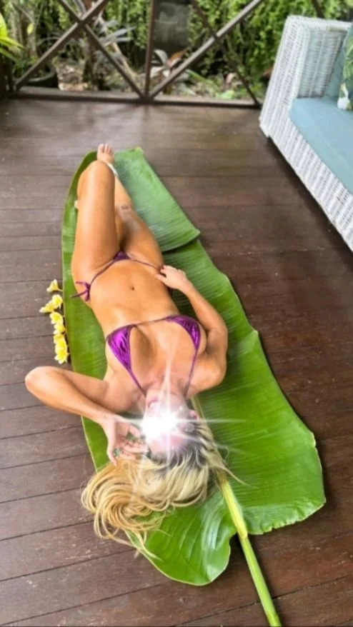A woman in a purple bikini lying on a large green leaf on a wooden porch, taking a selfie with her phone.