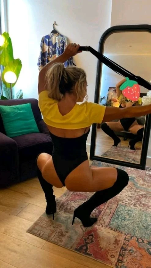 A woman in a black bodysuit, yellow crop top, and black thigh-high heels taking a mirror selfie in a living room. She is squatting with one knee on the floor surroundings include a purple couch, a green pillow, a large leafy plant, and a patterned rug.