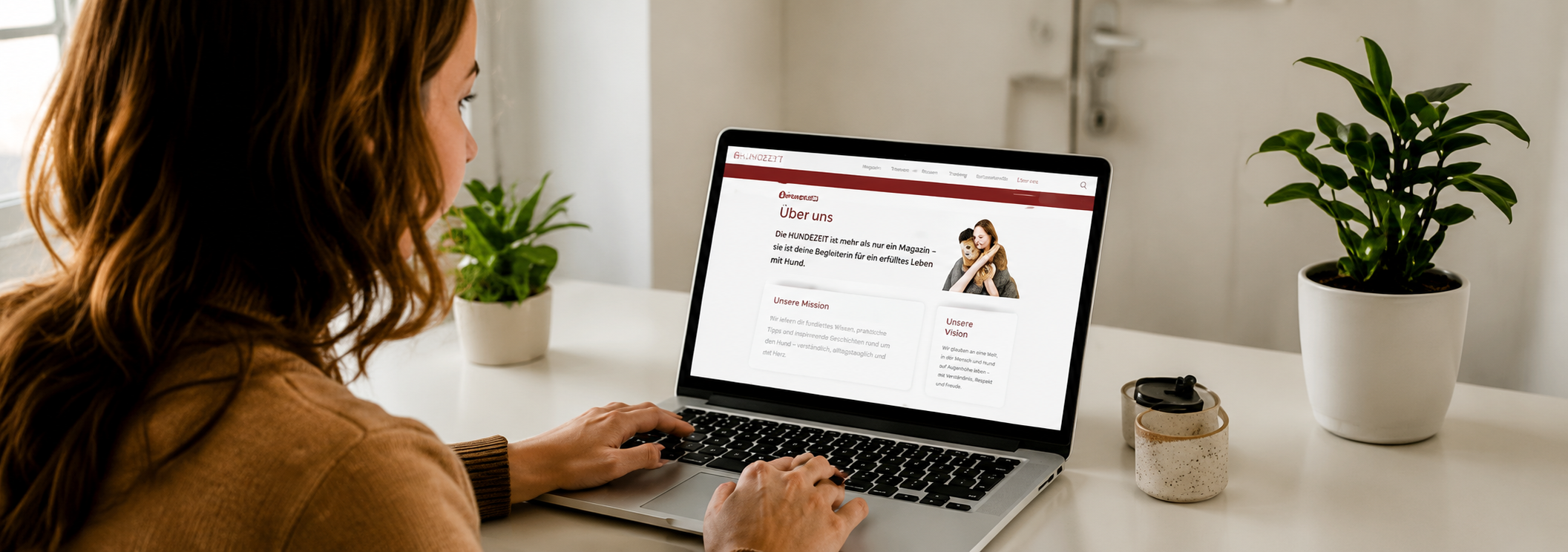 Eine Frau sitzt an einem weißen Tisch vor einem Laptop, der eine Webseite über Hunde zeigt. Der Tisch hat zwei kleine Pflanzentöpfe mit grünen Pflanzen, eine Tasse und ein schmales Gefäß. Die Frau hat braune, gewellte Haare und trägt einen braunen Pullover.