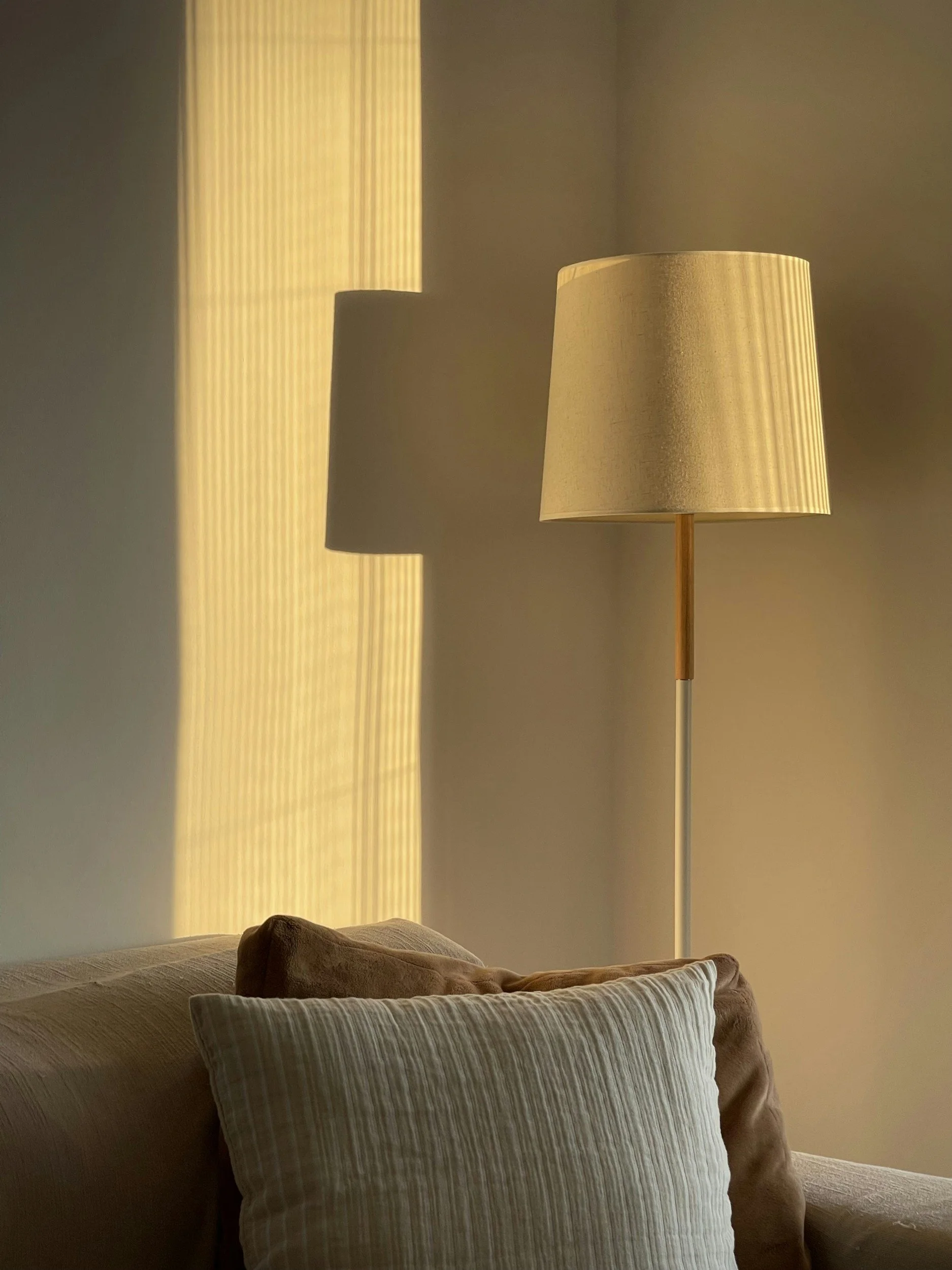 A cozy living room corner with a beige couch, striped cushion, standing lamp with a beige shade, and sunlight streaming through a window with vertical blinds, creating a shadow on the wall.