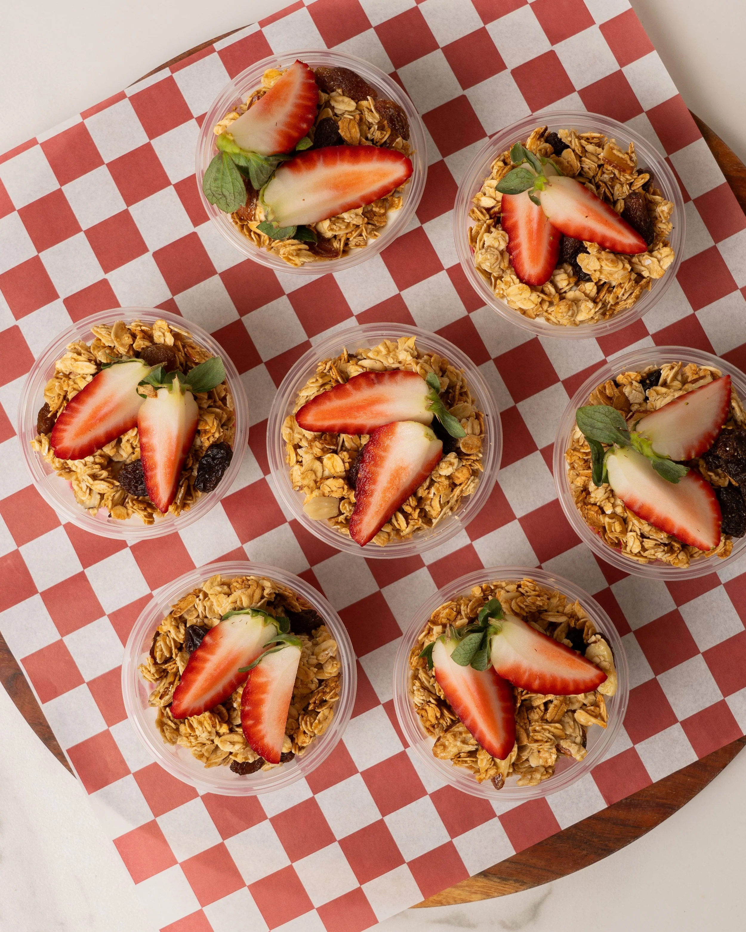 Multiple small cups of granola topped with sliced strawberries and fresh mint leaves, arranged on a checkered paper on a wooden serving tray.