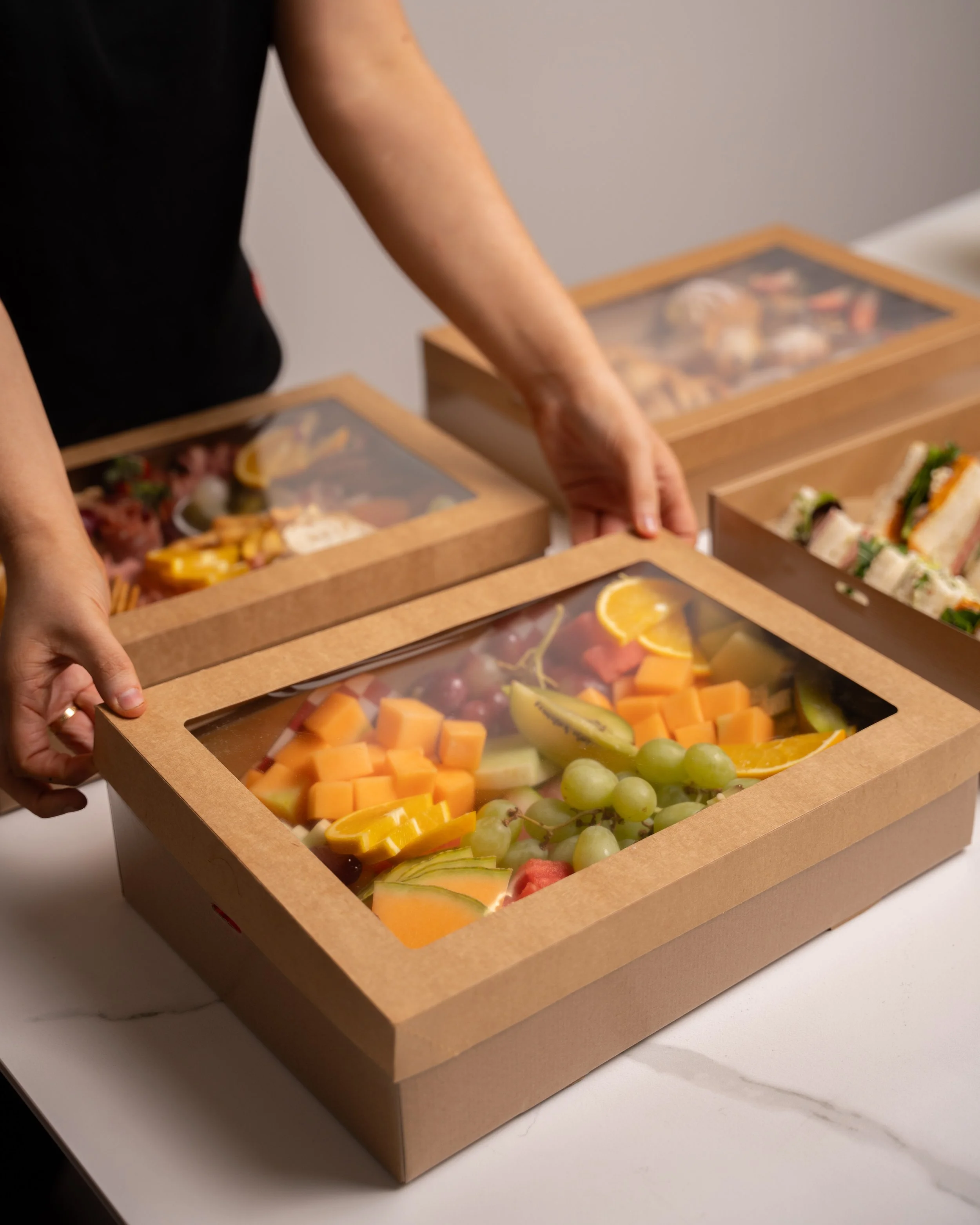 Close-up of a hand reaching into a wooden box with a transparent lid, containing an assortment of cut fruit including melon, grapes, and berries.