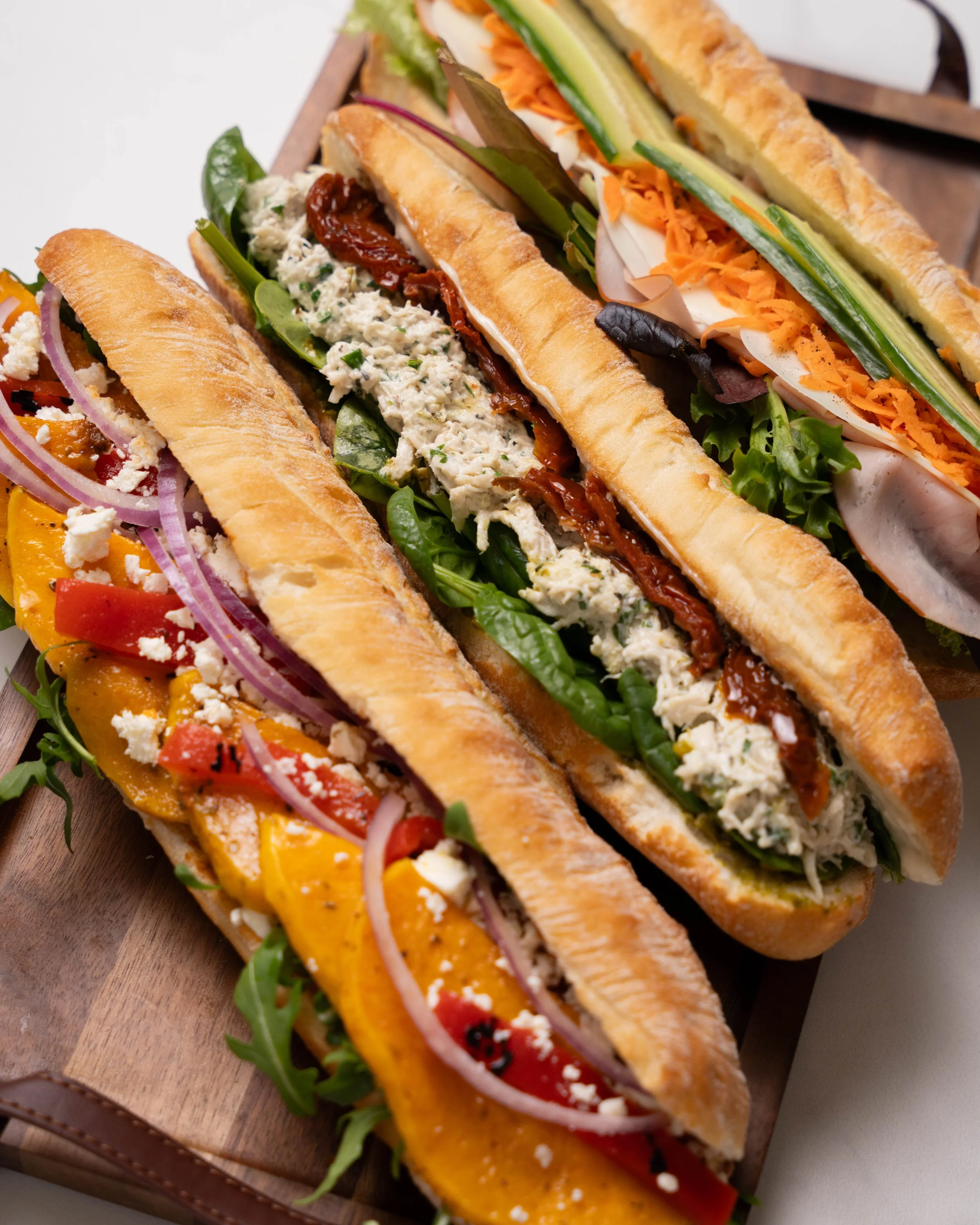 Three long cut sandwiches with various fillings including shredded cheese, sliced vegetables, greens, and sauces on a wooden serving board.