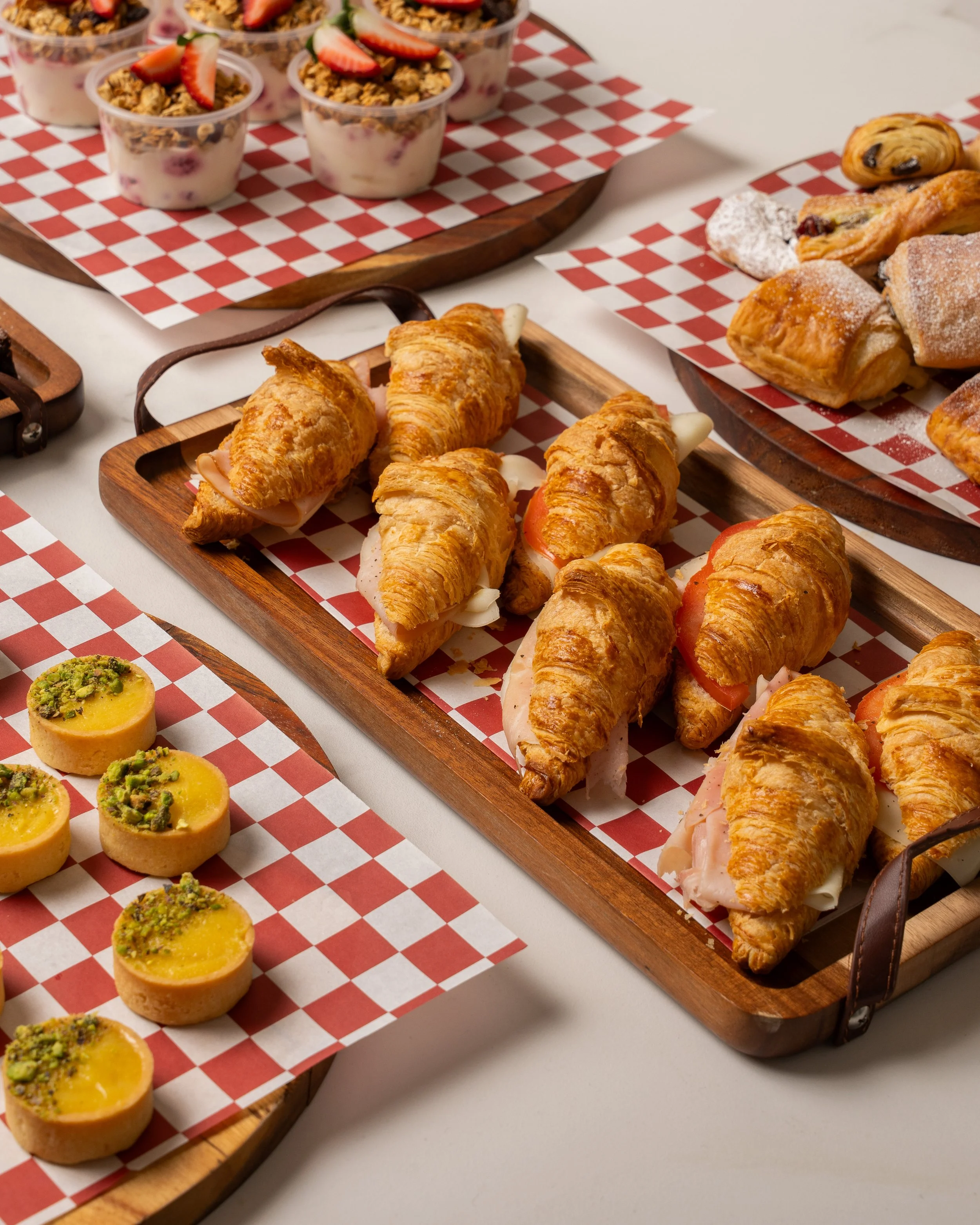 Assorted baked goods including mini lemon tarts topped with pistachios, croissant-wrapped sandwiches with turkey, ham, cheese, tomato, and lettuce, and various pastries with powdered sugar, on wooden serving trays lined with checkered paper.