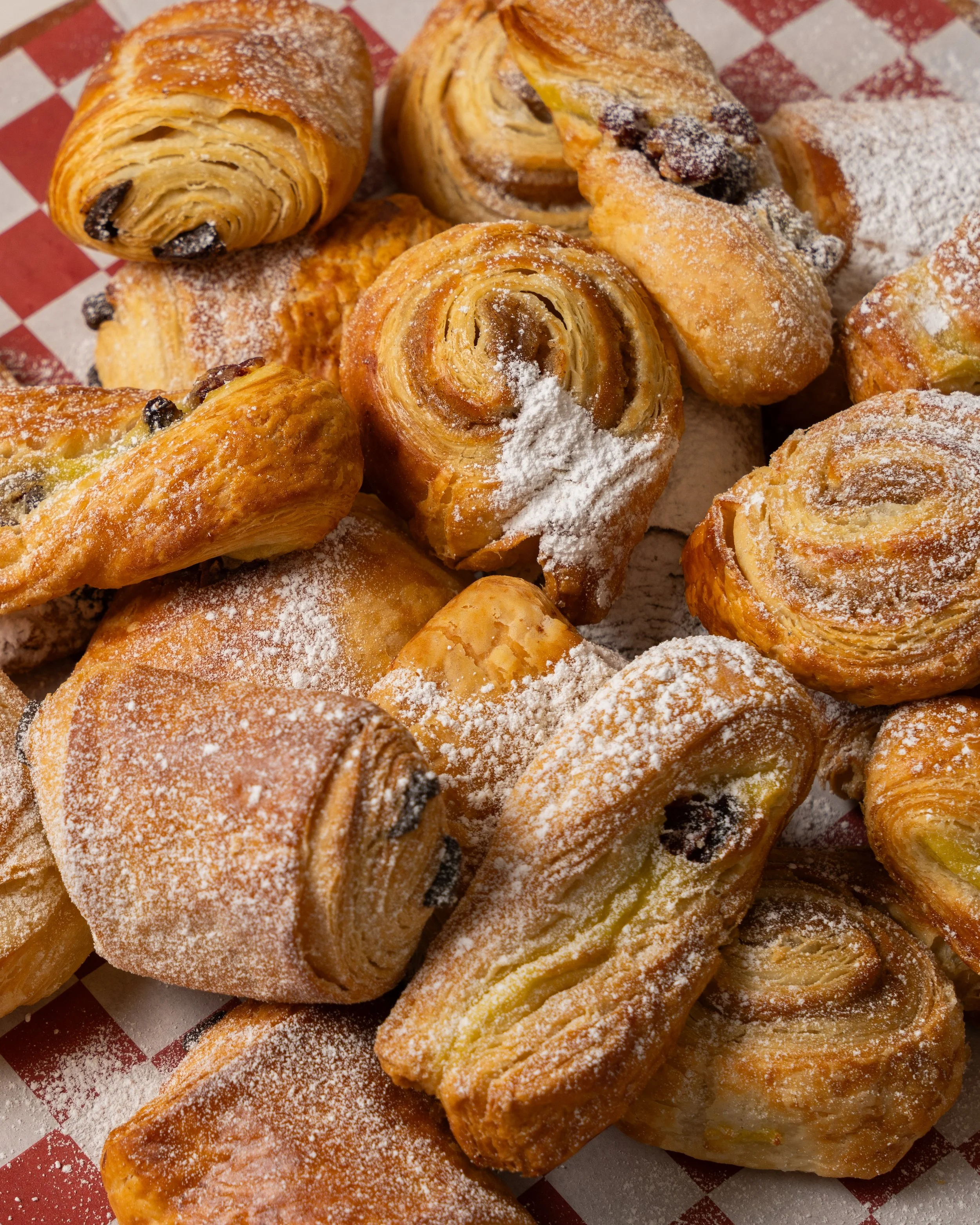 Assorted Danish pastries dusted with powdered sugar on red and white checkered paper.