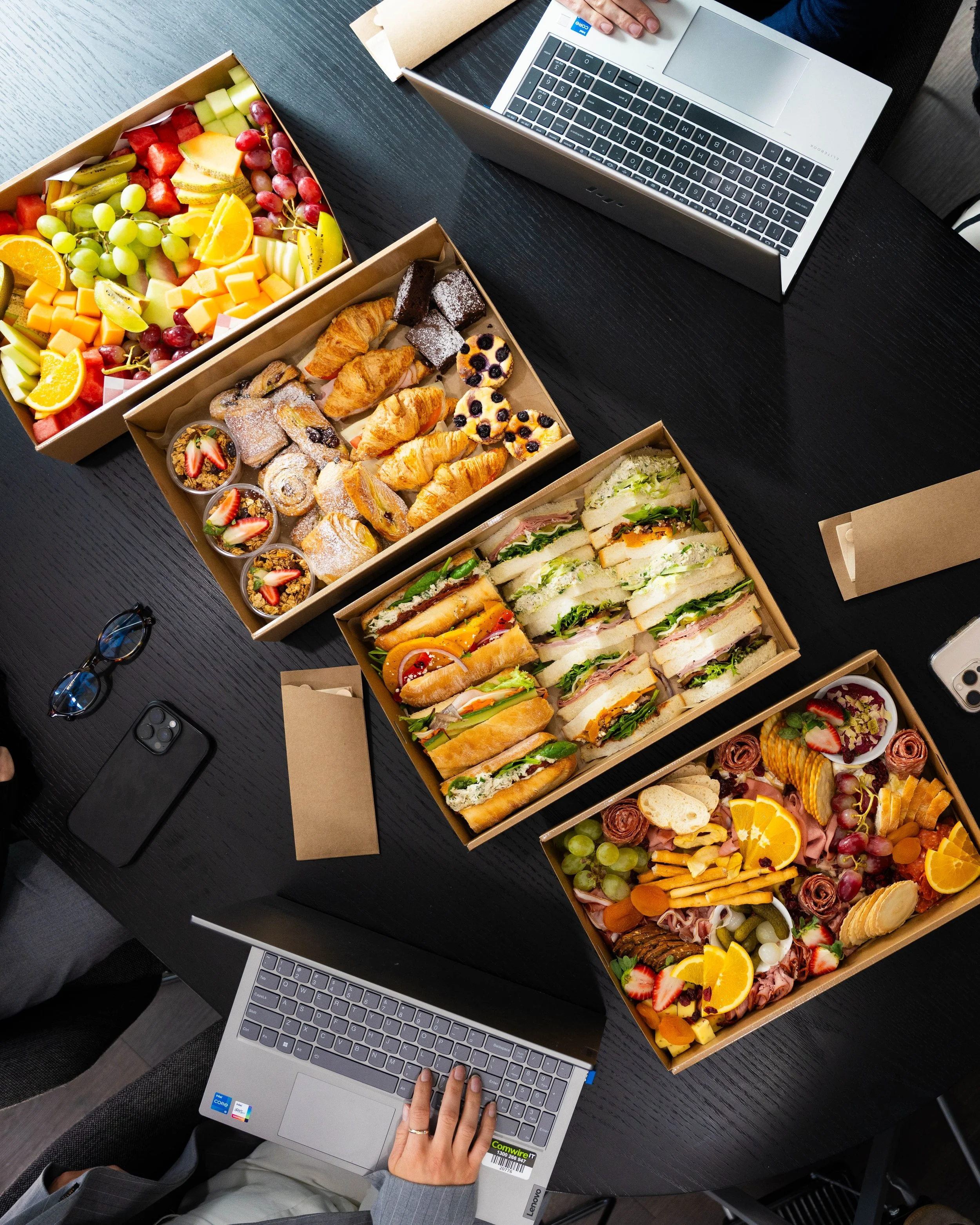 A top-down view of a workspace showing a person using a laptop, surrounded by several boxes of assorted food items including a large fruit platter, pastries, sandwiches, and desserts, on a black table.