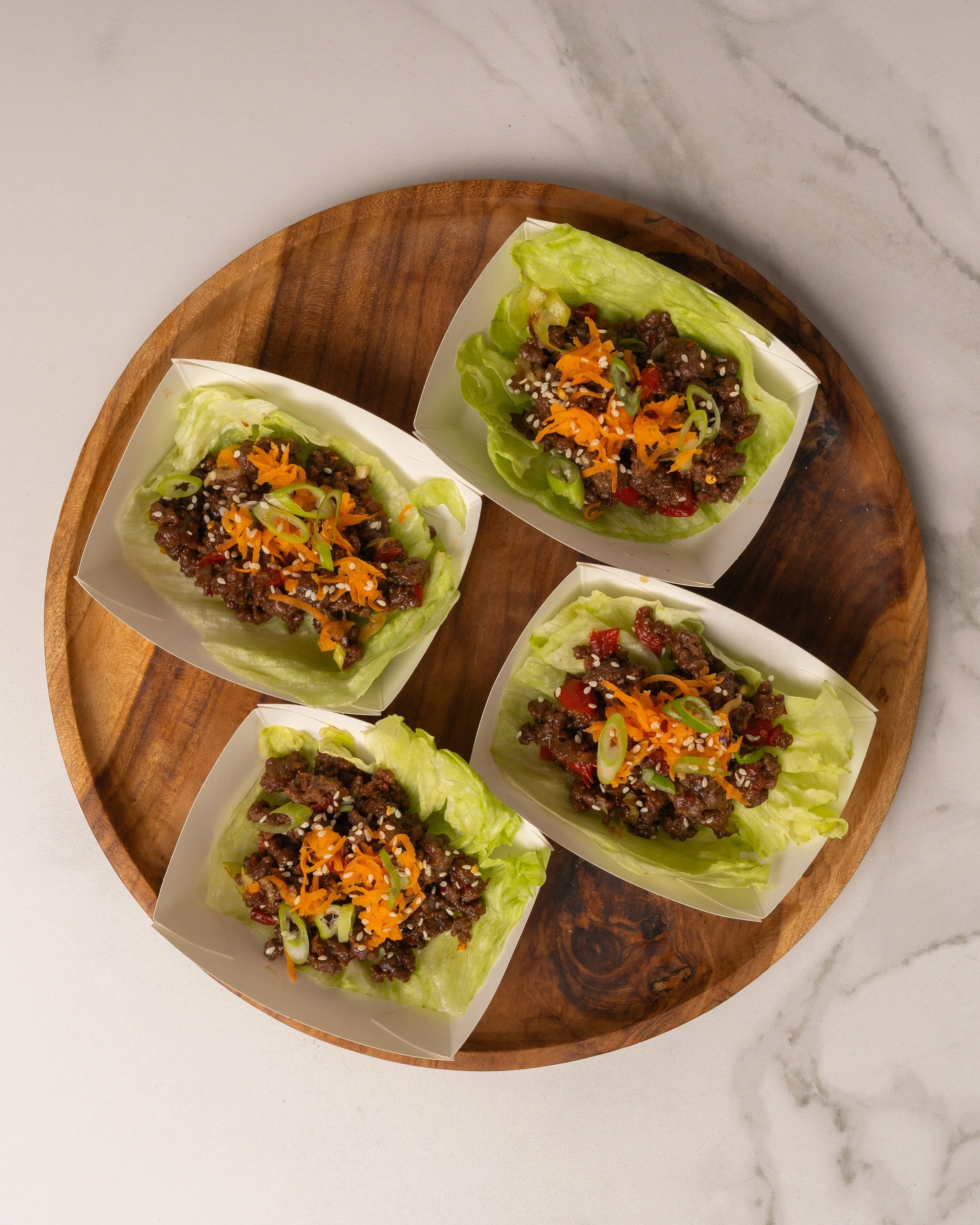 Four lettuce wraps filled with seasoned ground beef, shredded cheese, chopped green onions, and diced red peppers on a round wooden platter.