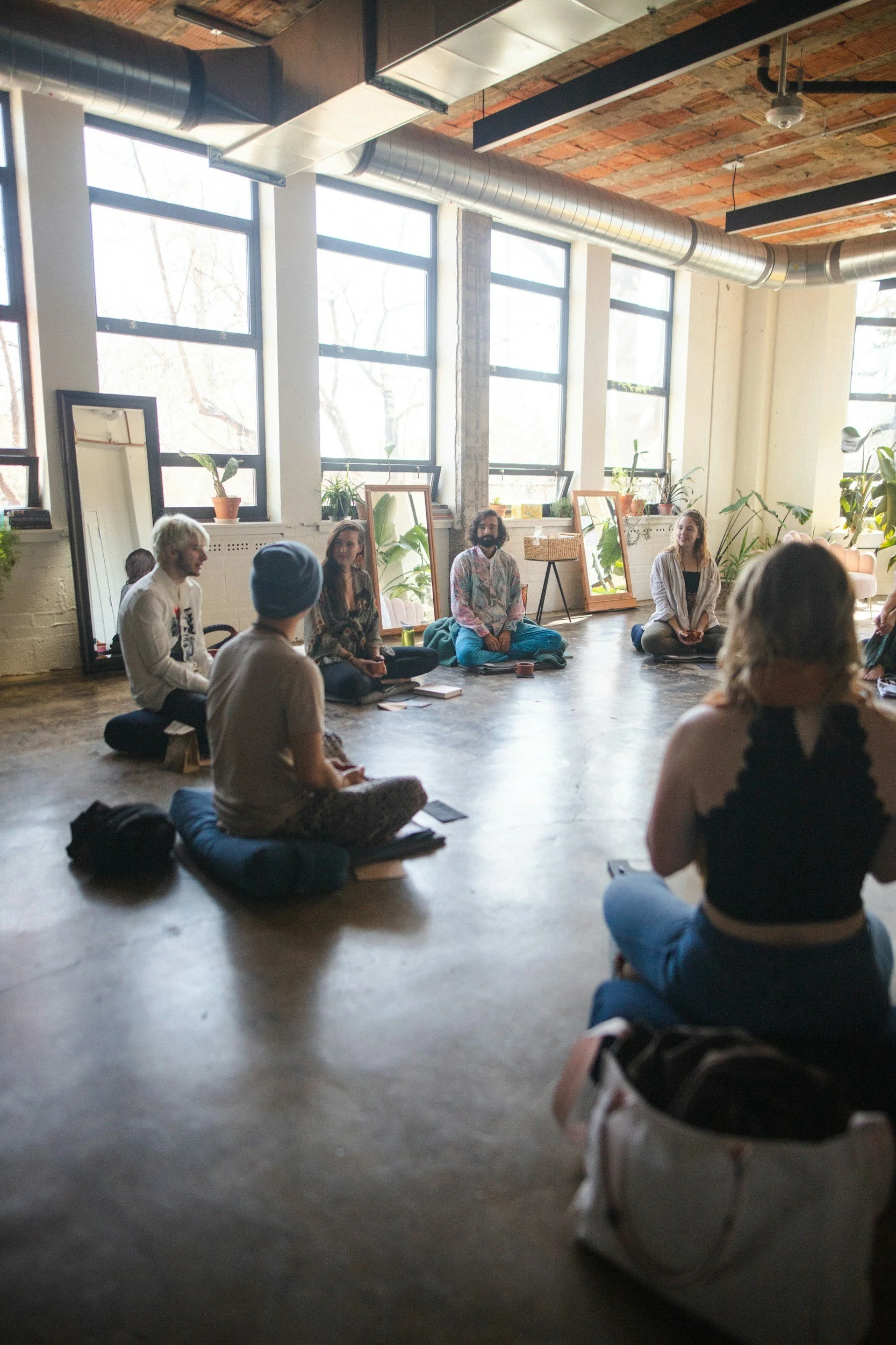 Group of people sitting in a circle on the floor of a brightly lit room with large windows, plants, and minimalistic decor.