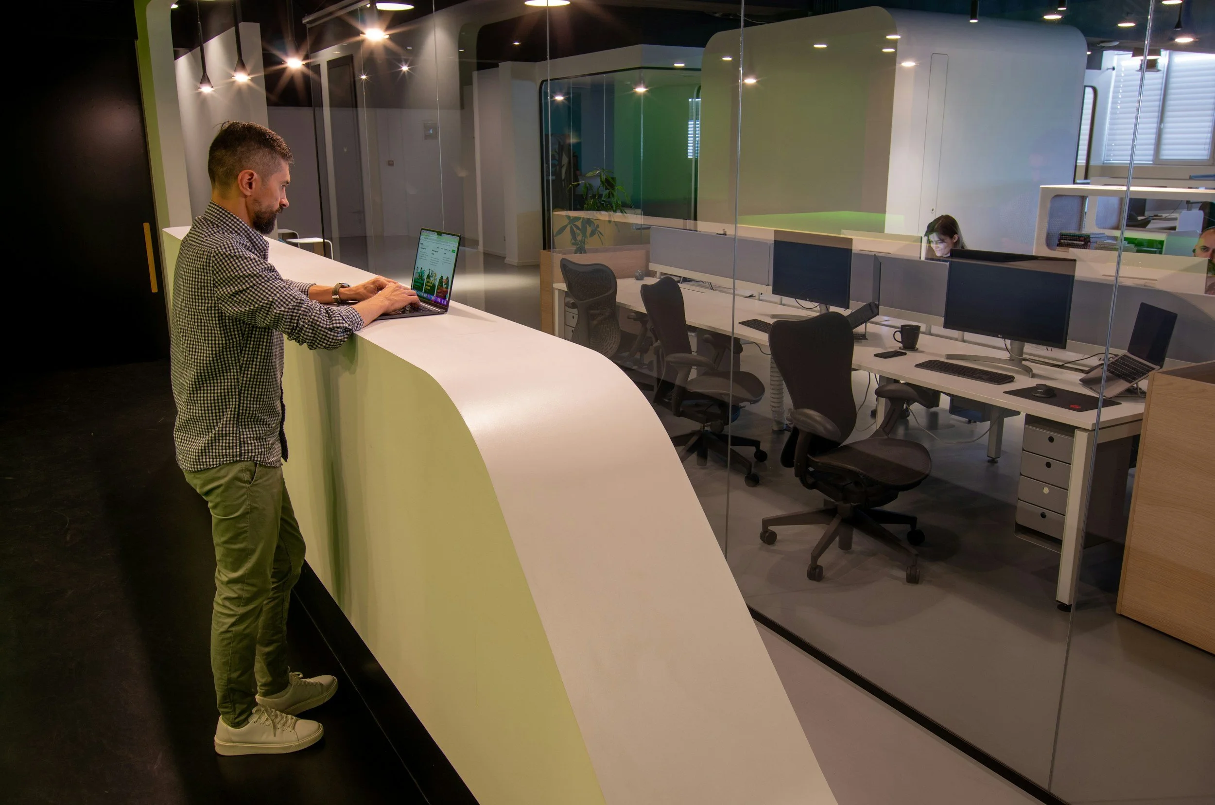 Man standing at a white reception desk, working on a laptop in a modern office with glass-walled cubicles and employees working at desks inside.