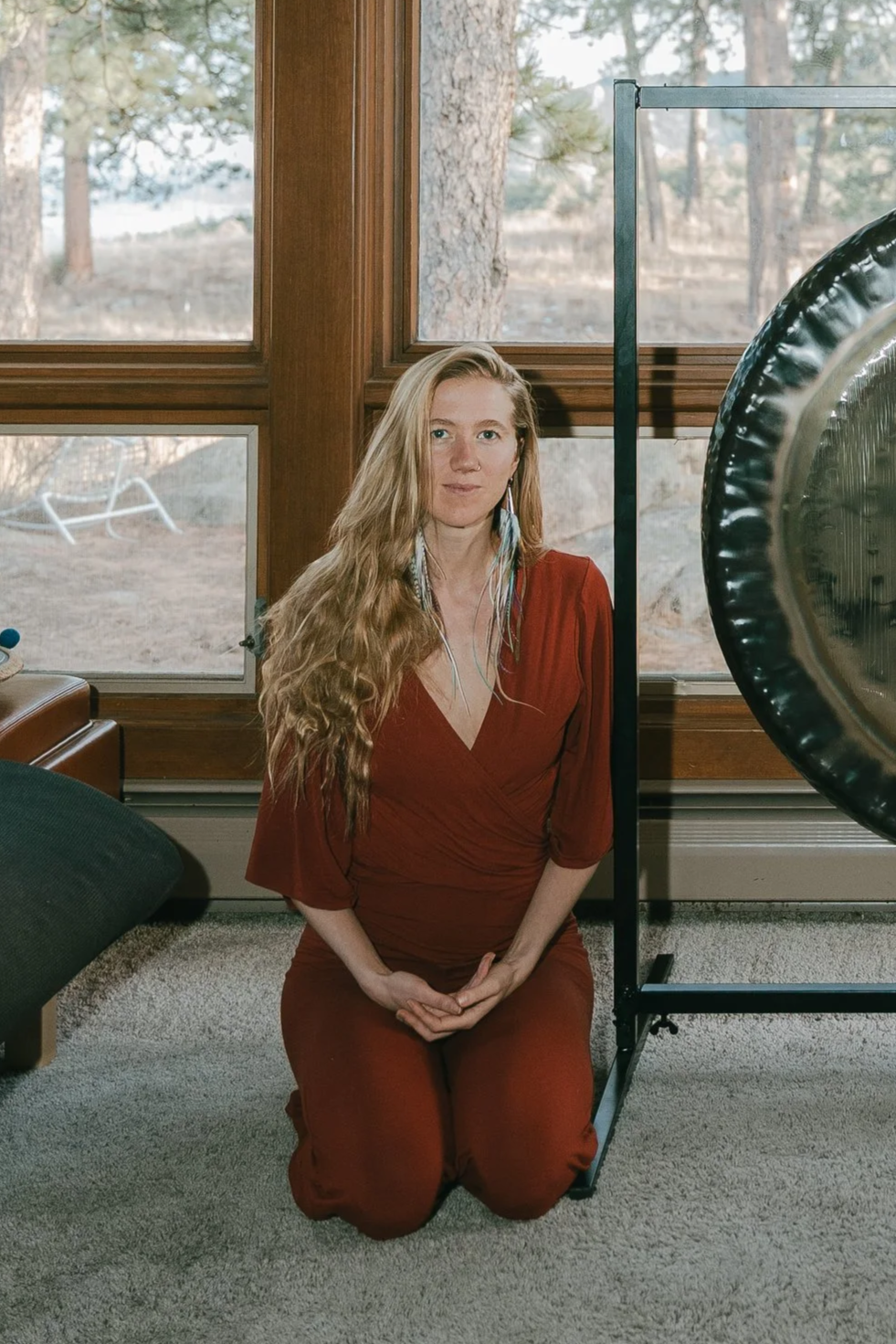 A woman with long wavy blonde hair kneeling inside near a large window, wearing a red outfit, with a gong on a stand beside her.