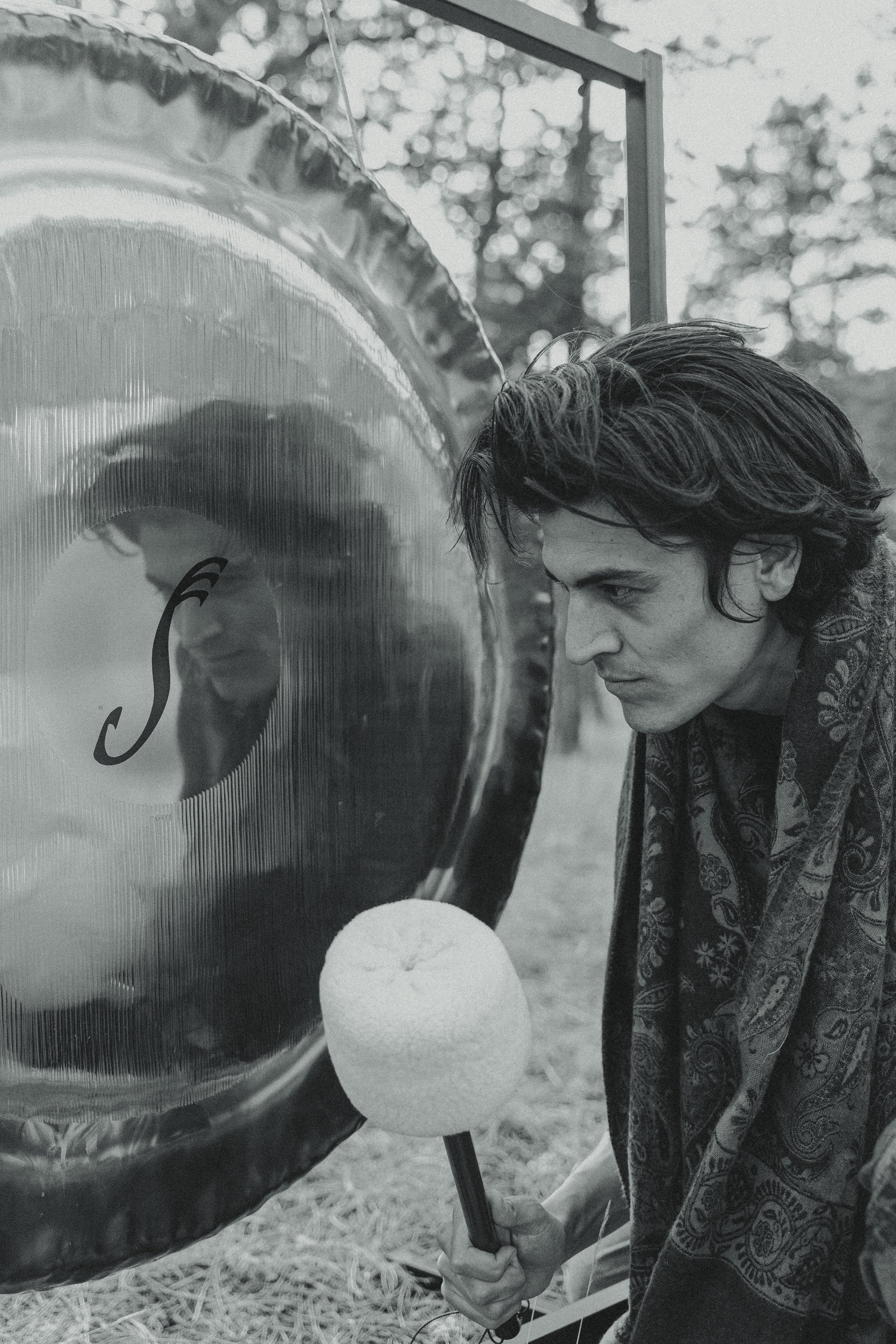 A young man with dark, wavy hair and a patterned scarf leans forward with intense focus while about to strike a gong with a mallet, outdoors with trees in the background.