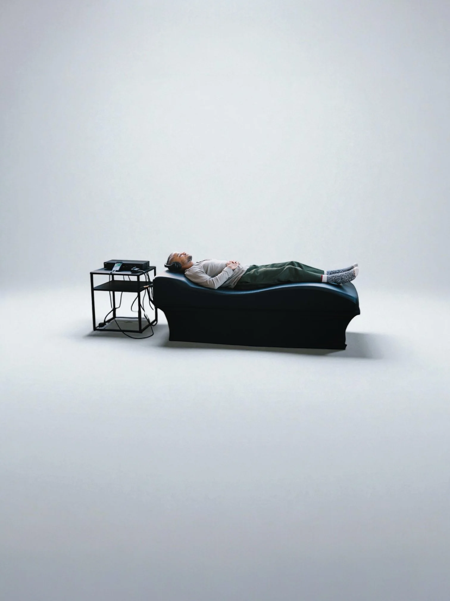 Person lying on a black lounge chair listening to music with headphones, next to a small black table with electronic devices, in a minimalistic white space.