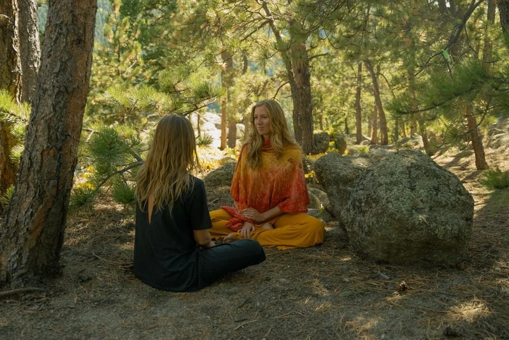 Two women sitting cross-legged on the ground in a forest, facing each other. One is wearing black, the other is dressed in a colorful, orange and yellow outfit, and they appear to be engaged in a conversation.