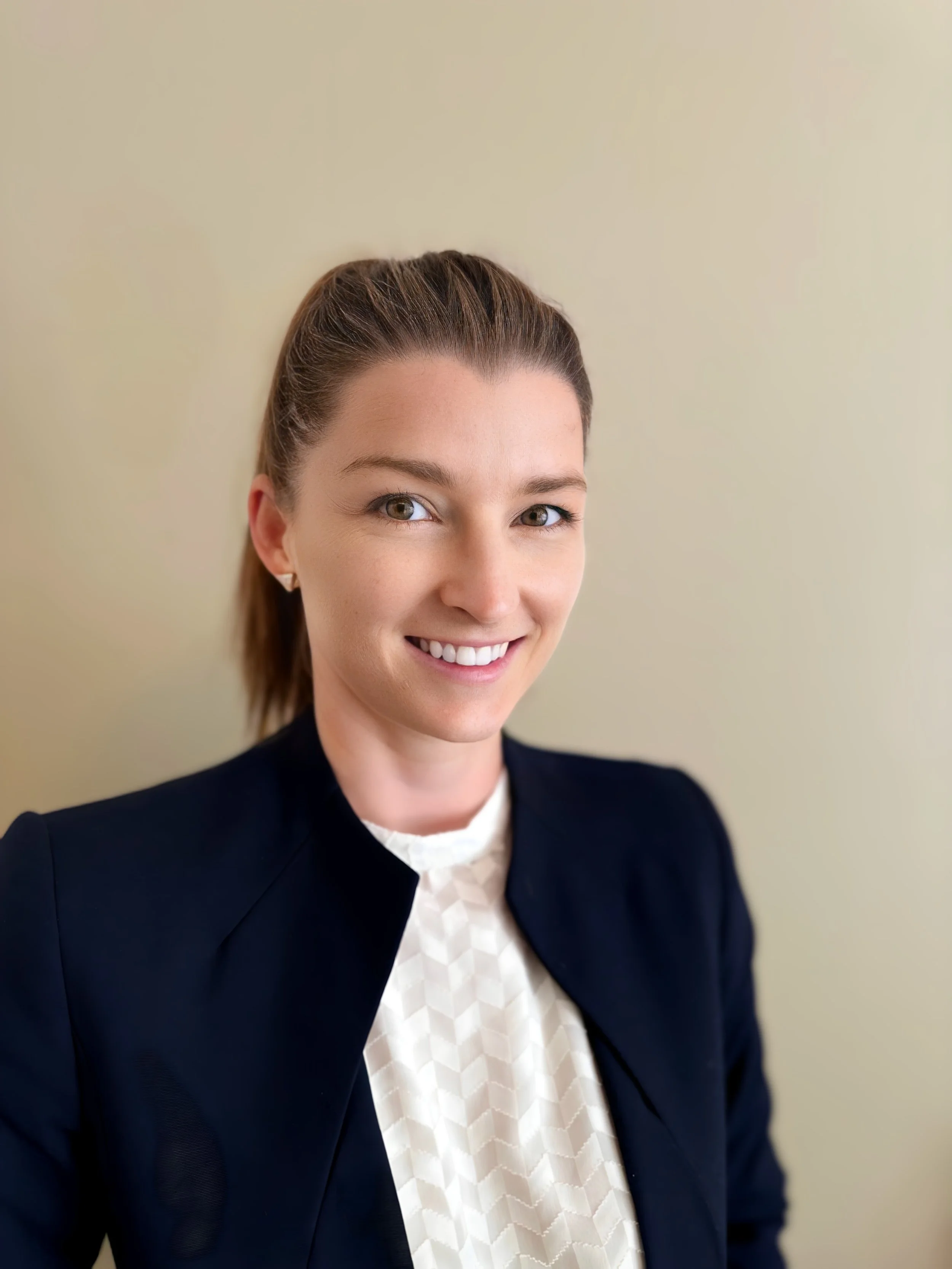 Professional woman with brown hair in a ponytail, smiling, wearing a black blazer and white patterned blouse, standing in front of a plain beige wall.