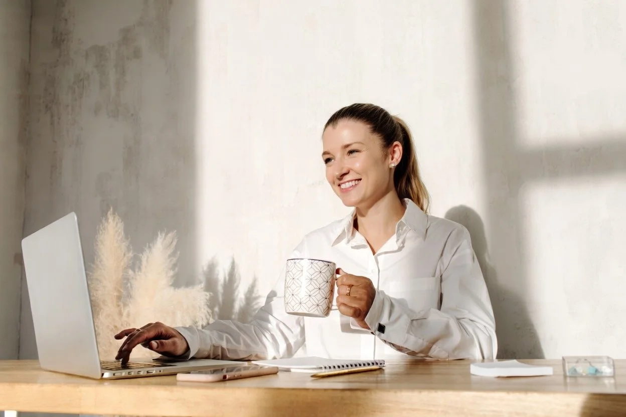 A woman sitting at a desk, smiling, holding a coffee mug in her left hand, with a laptop in front of her.