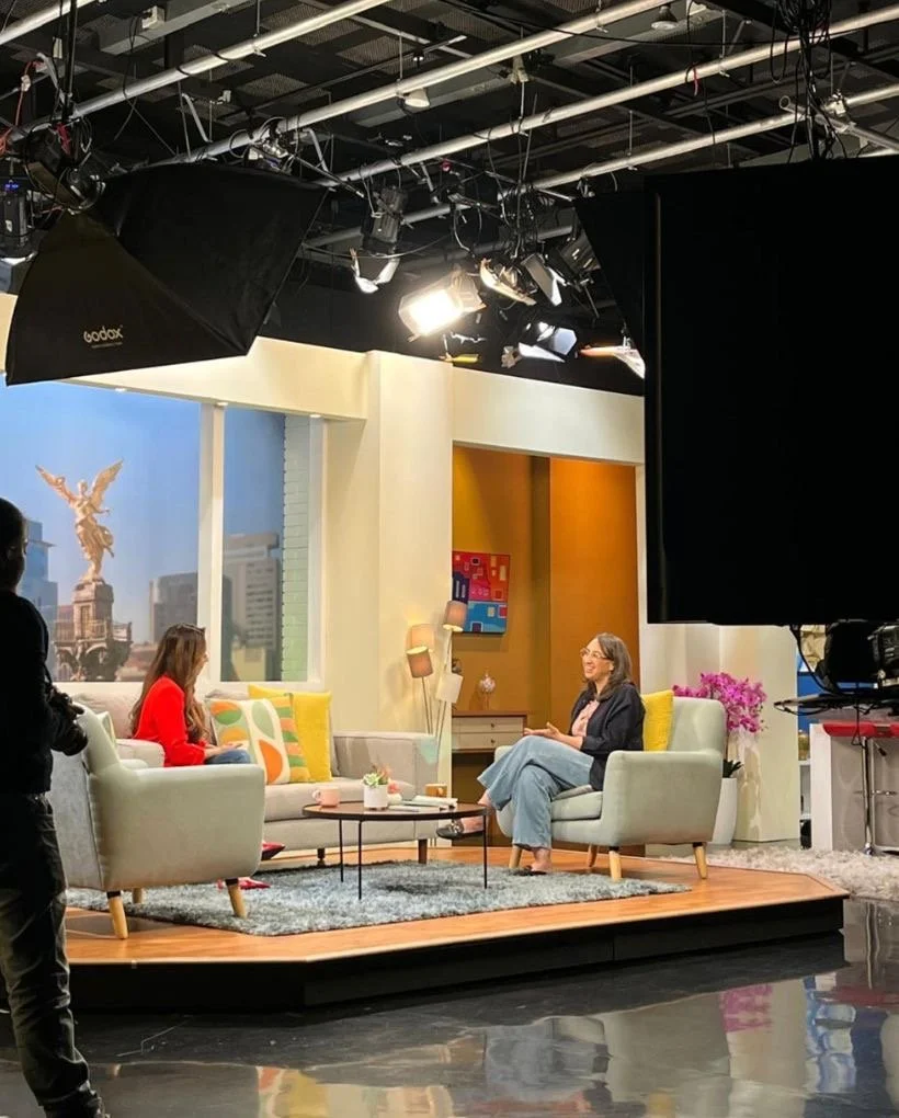 Cynthia Ramírez talking on a television set with armchairs and colorful decorations, with recording equipment and lights overhead.