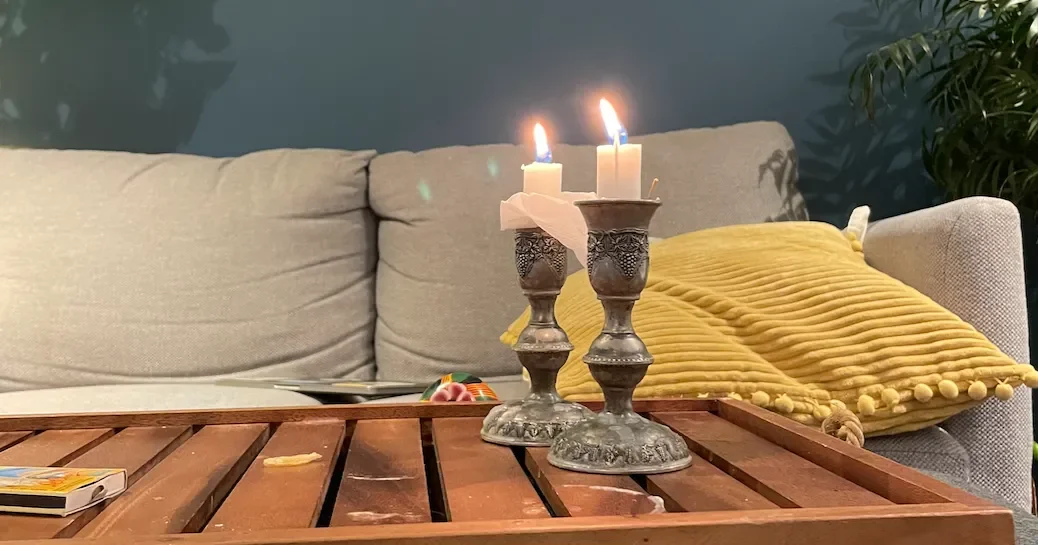 A lit candle in metal candleholders for Shabbat on a wooden table in a room with a gray armchair and yellow cushions.