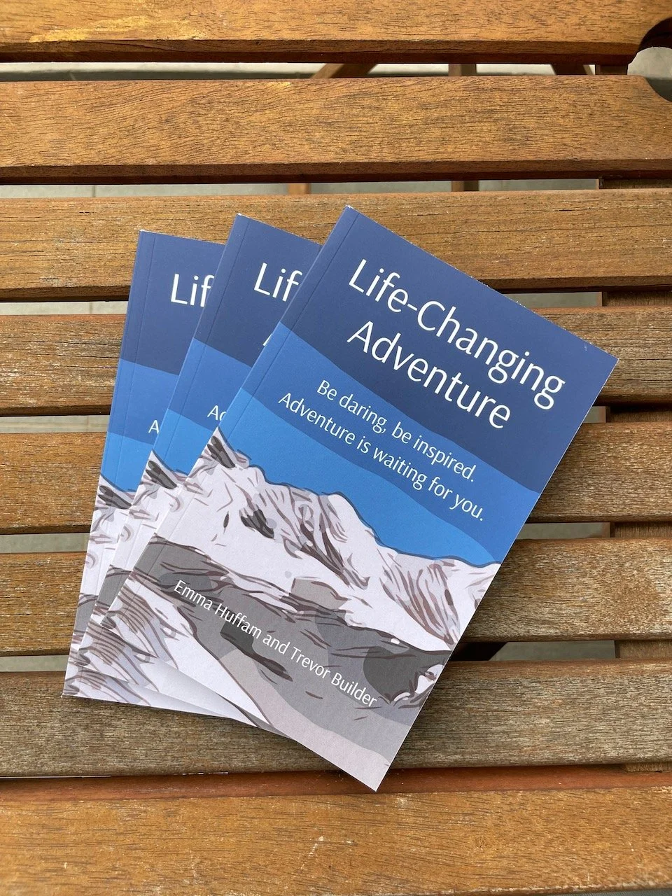 a wooden table upon which is 3 copies of the book by the Project Base8000 team called Life-Changing Adventure