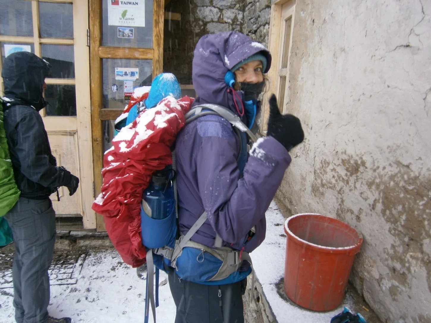 a trekker dressed in warm waterproof trekking gear with snow on backpack and giving the thumbs up
